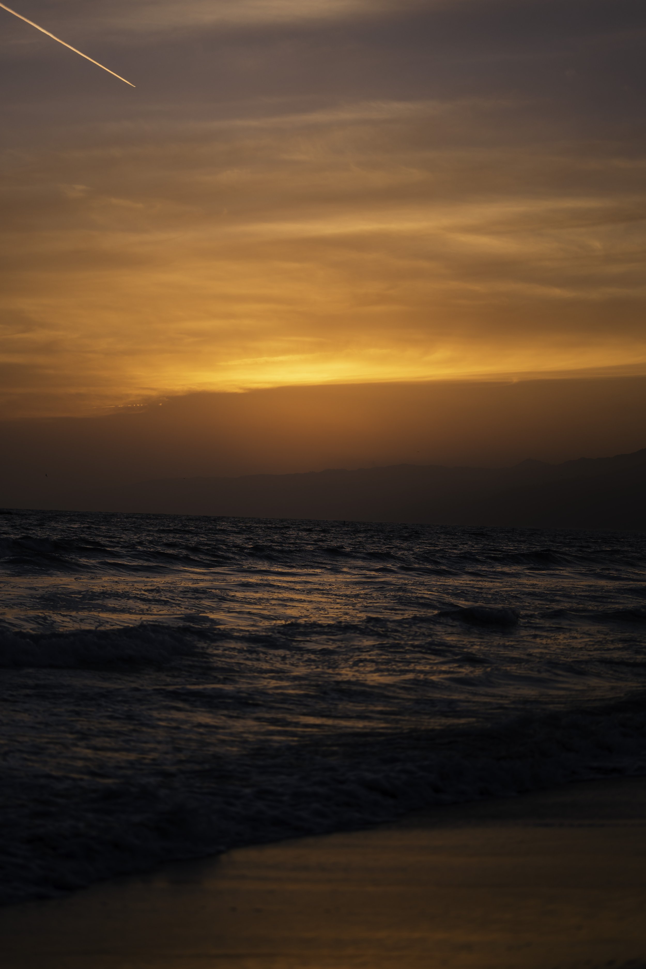 Sunset over ocean waves with a golden sky and a contrail.