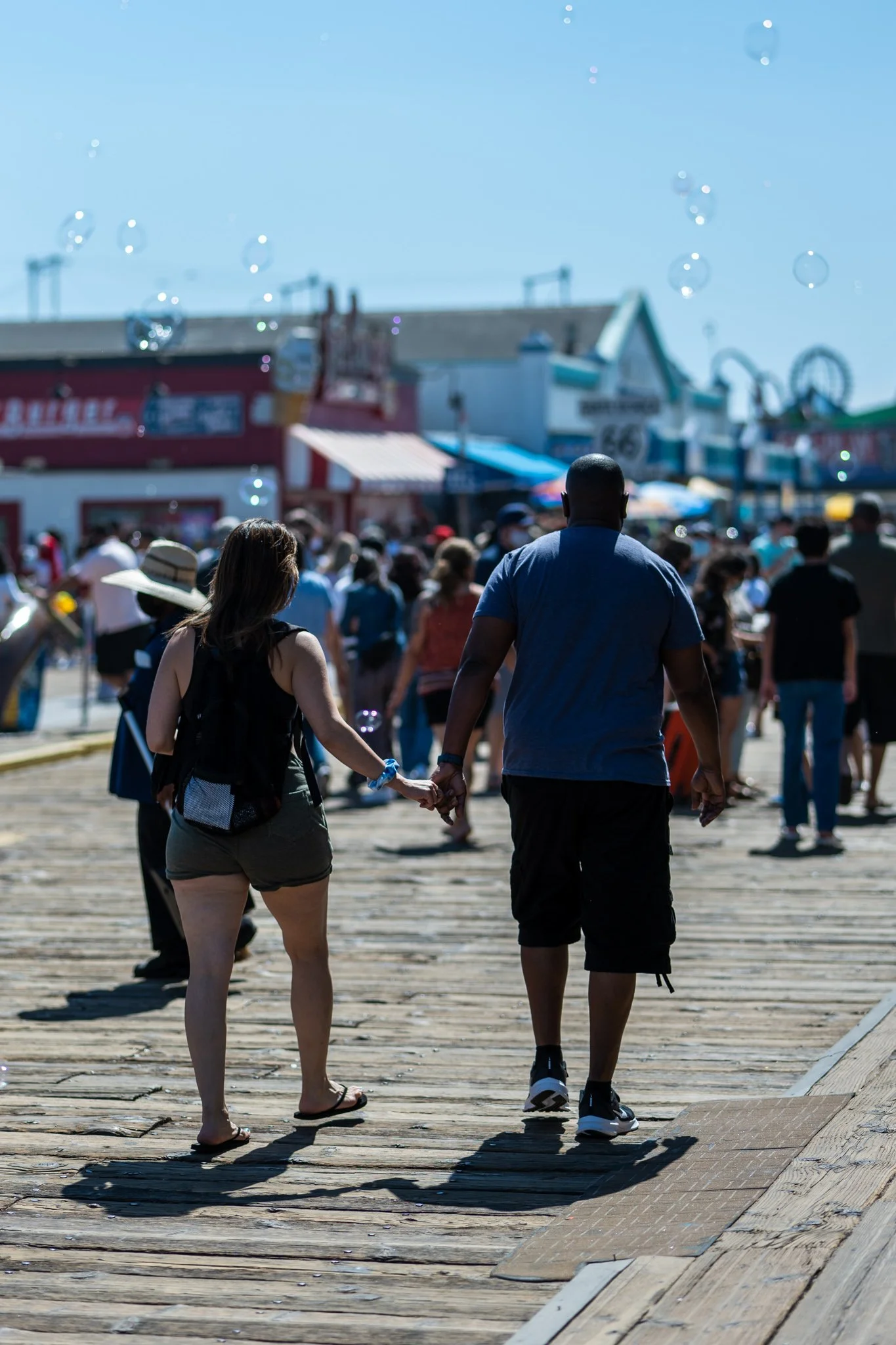 Couple holding hands walking on a crowded boardwalk with bubbles in the air, surrounded by people and buildings.