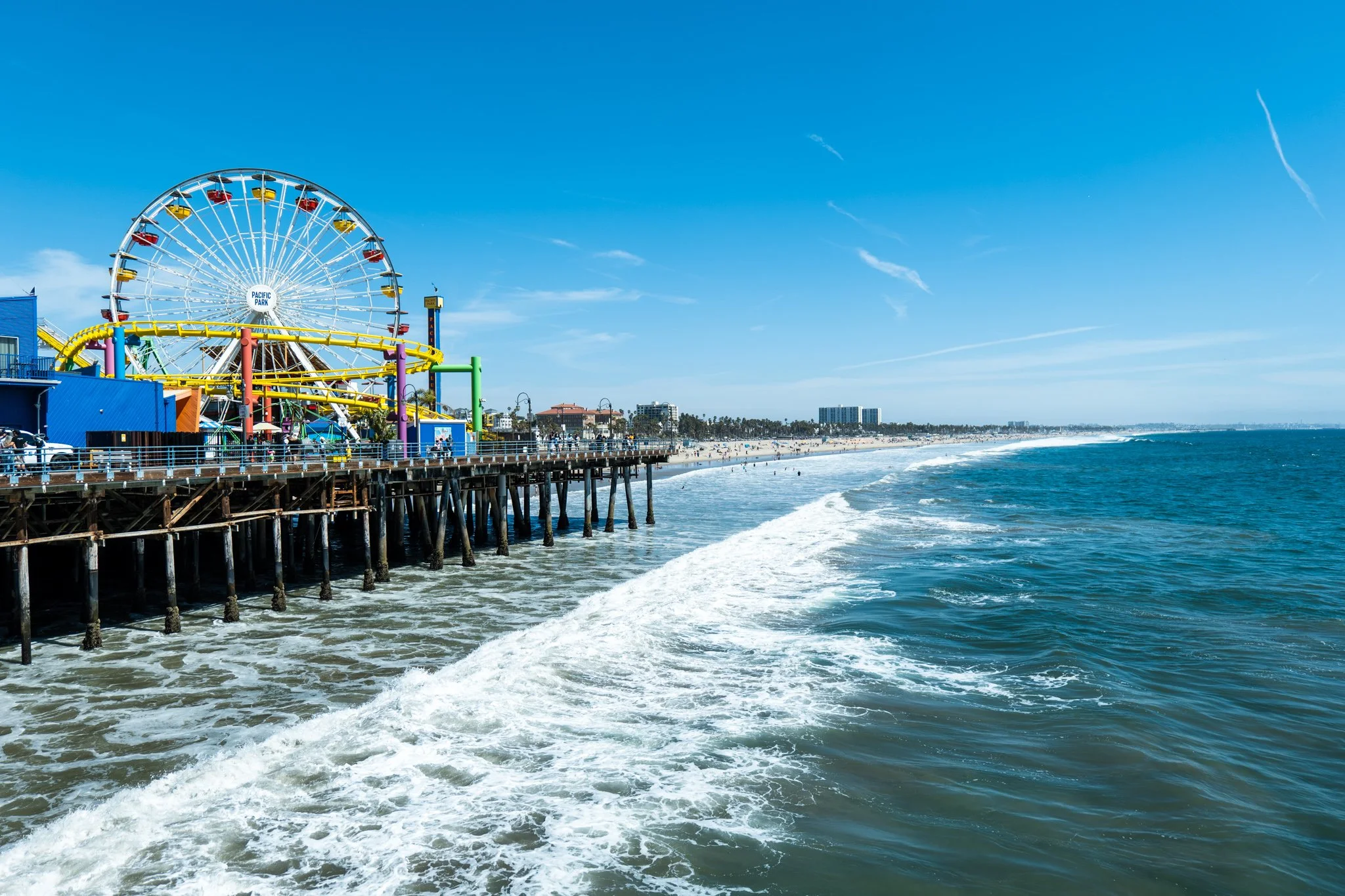 Santa Monica Pier with Ferris wheel and roller coaster, ocean waves, and beach in sunny weather.