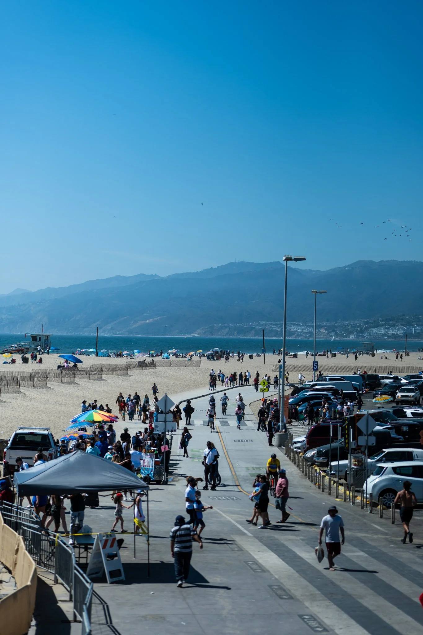 Crowded beach boardwalk with people walking, biking, and visiting vendors under a clear blue sky. Cars are parked nearby, and mountains are visible in the background.