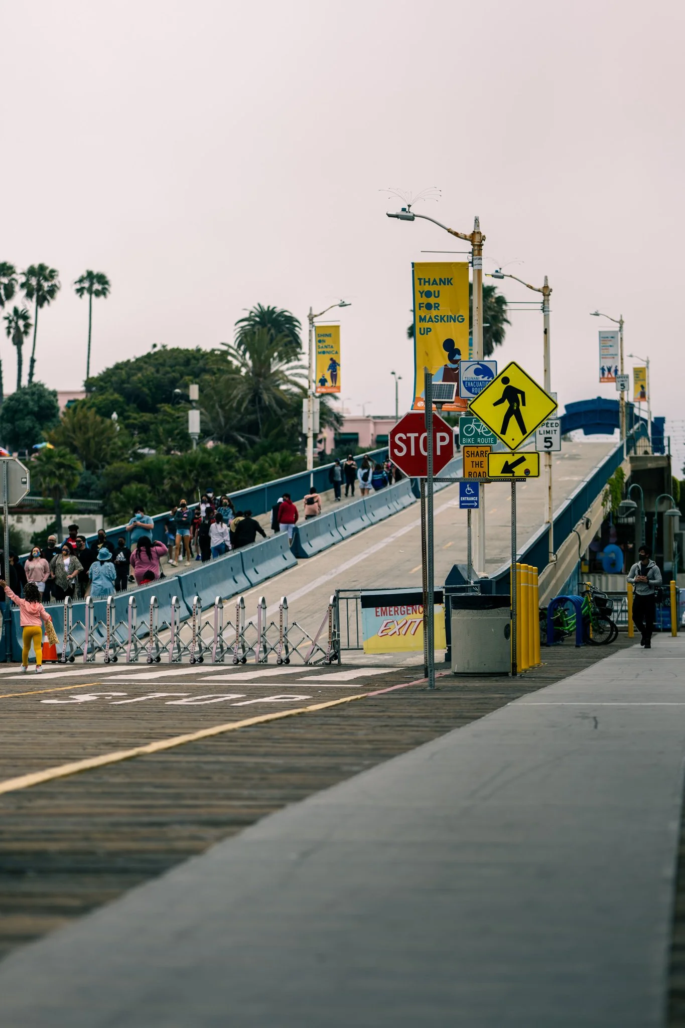 People walking on an elevated path with palm trees in the background, various signs including "Stop" and "Emergency Exit." A banner reads "Thank You For Masking Up."