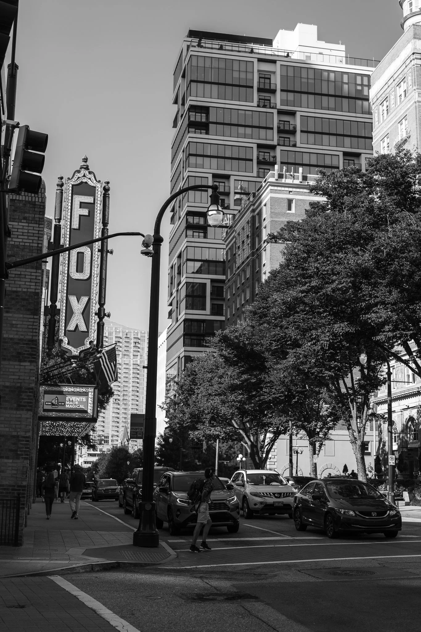 Black and white city street with "FOX" theater sign, cars, and people walking, surrounded by modern buildings and trees.