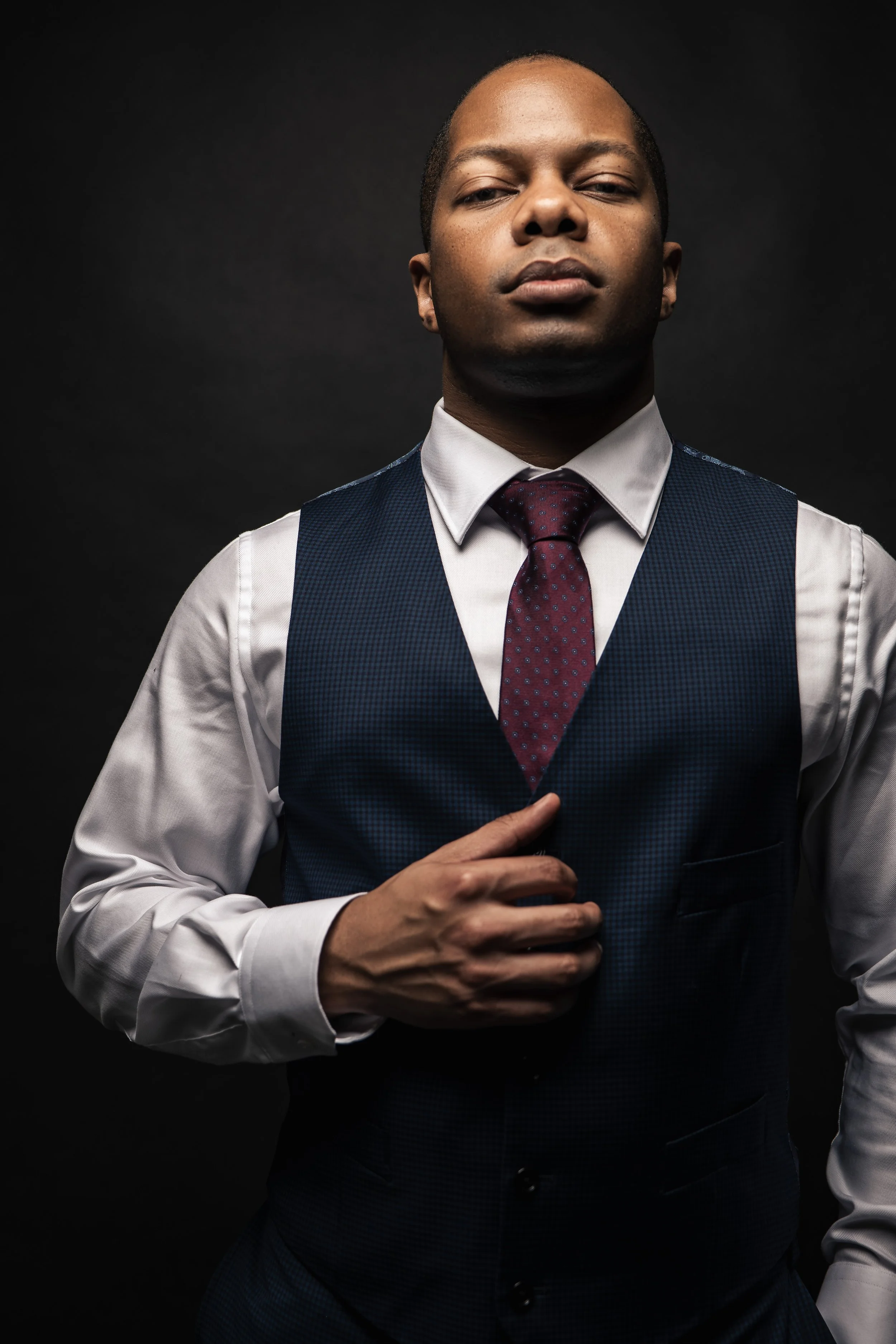 Man in a suit with dark background, wearing a white shirt and blue vest, adjusting a red tie, looking confident.