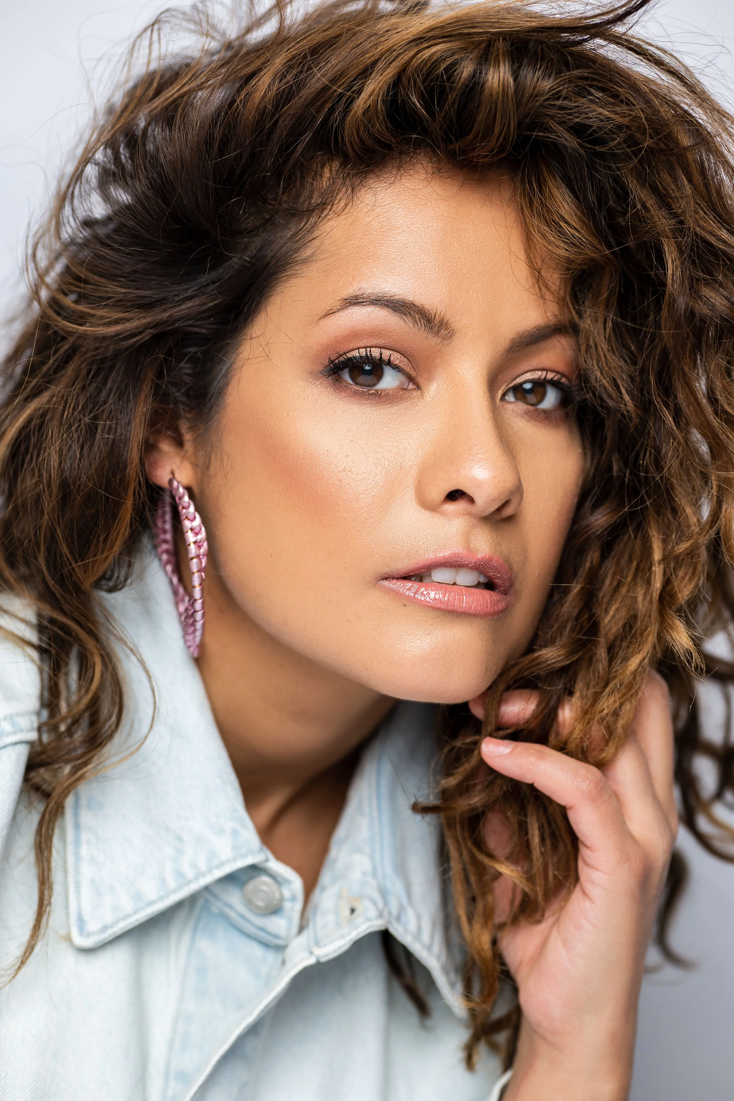 Woman with wavy hair, wearing a light denim jacket and large pink hoop earrings.