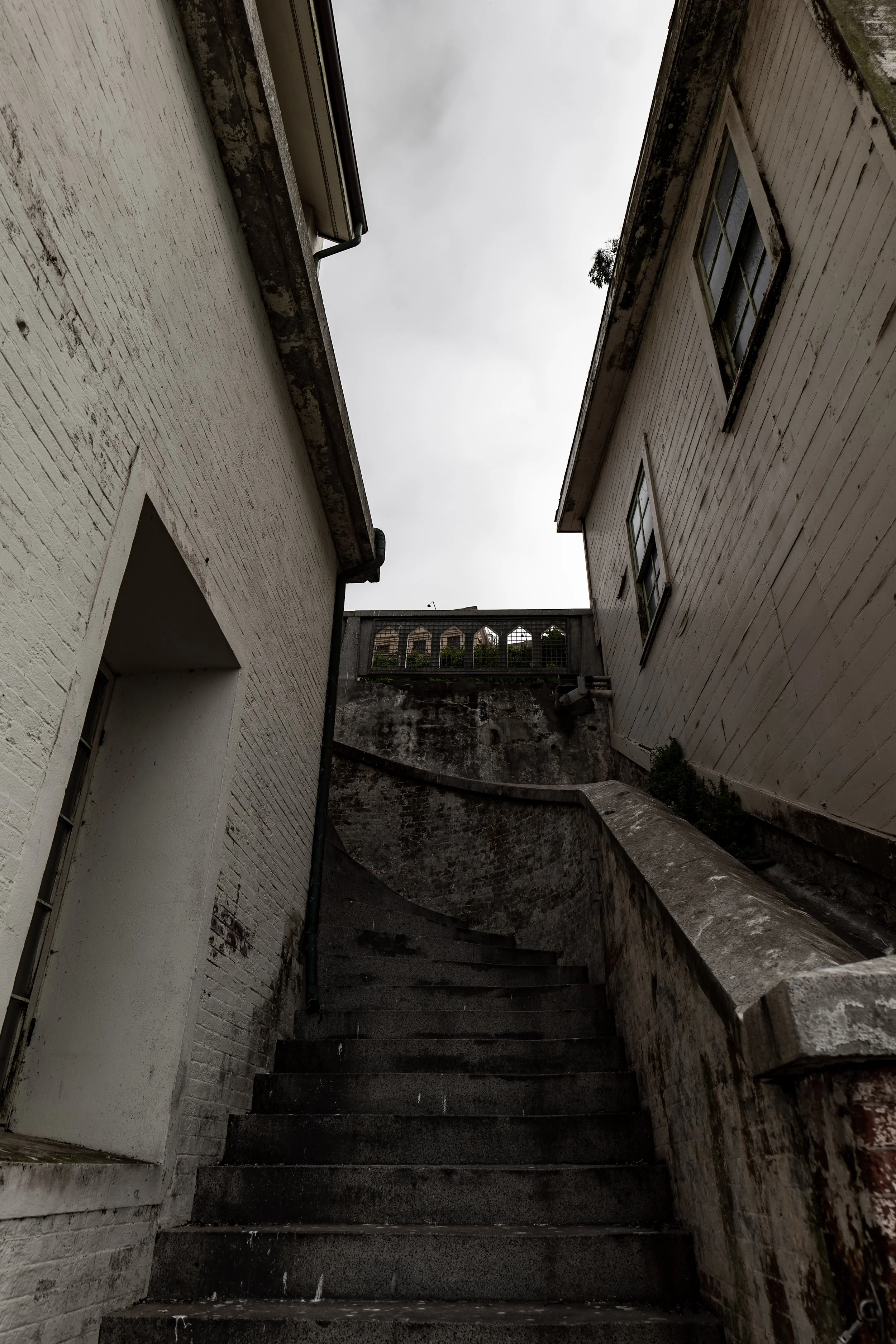 Narrow stairway between two old brick buildings, leading upward to a small arched fence at the top. Cloudy sky above.