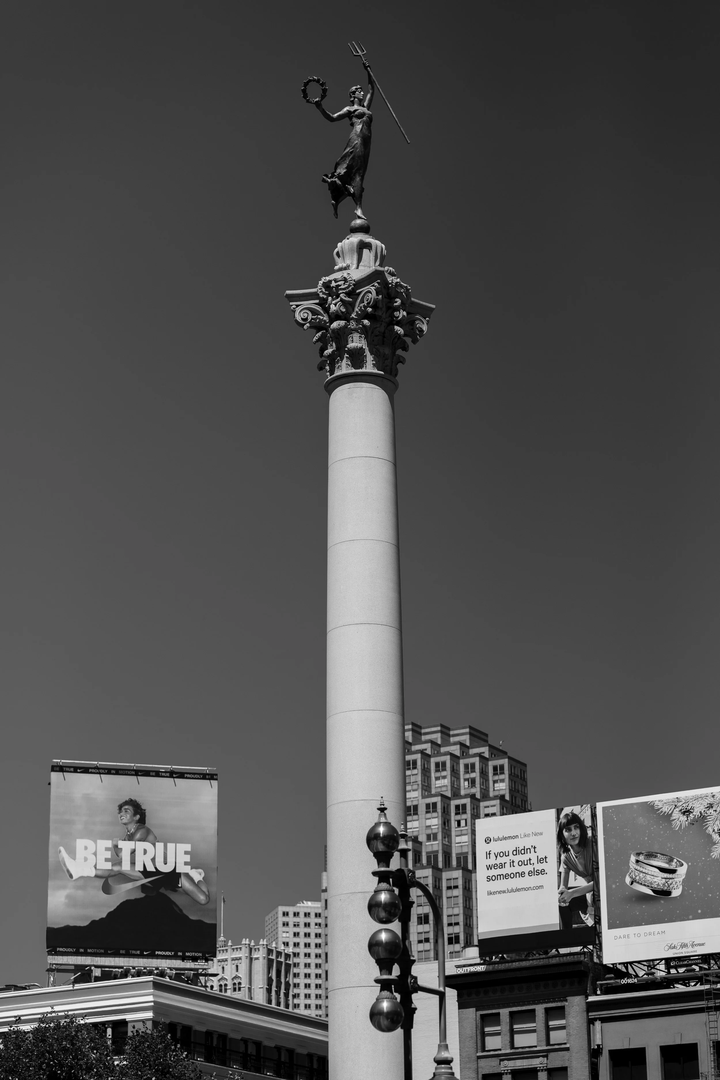 Black and white photo of a tall column with a statue on top, holding a trident and wreath. Behind are billboards and city buildings.