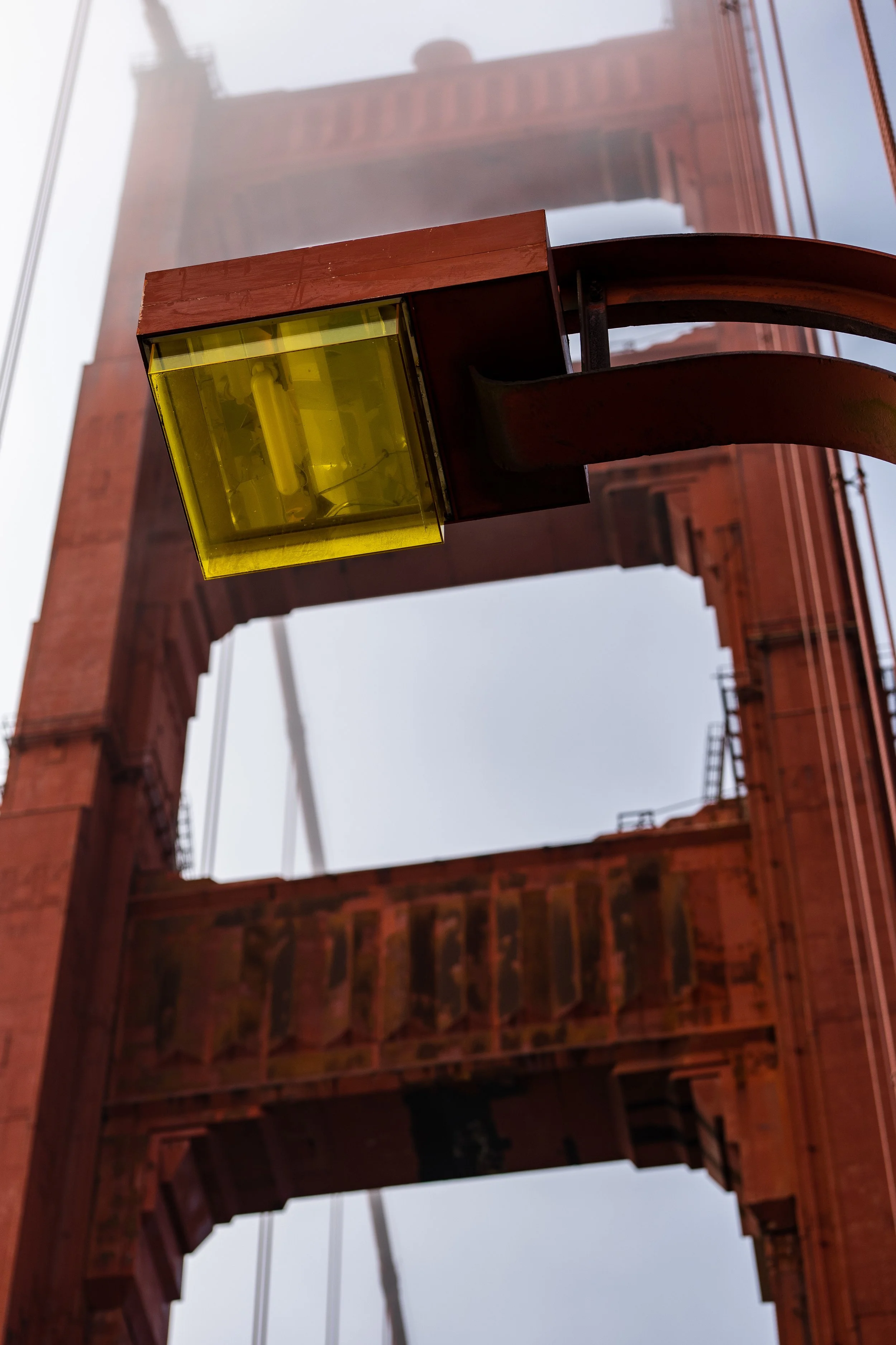 Close-up of a yellow light fixture with the Golden Gate Bridge tower and cables visible in the background, partially covered by fog.