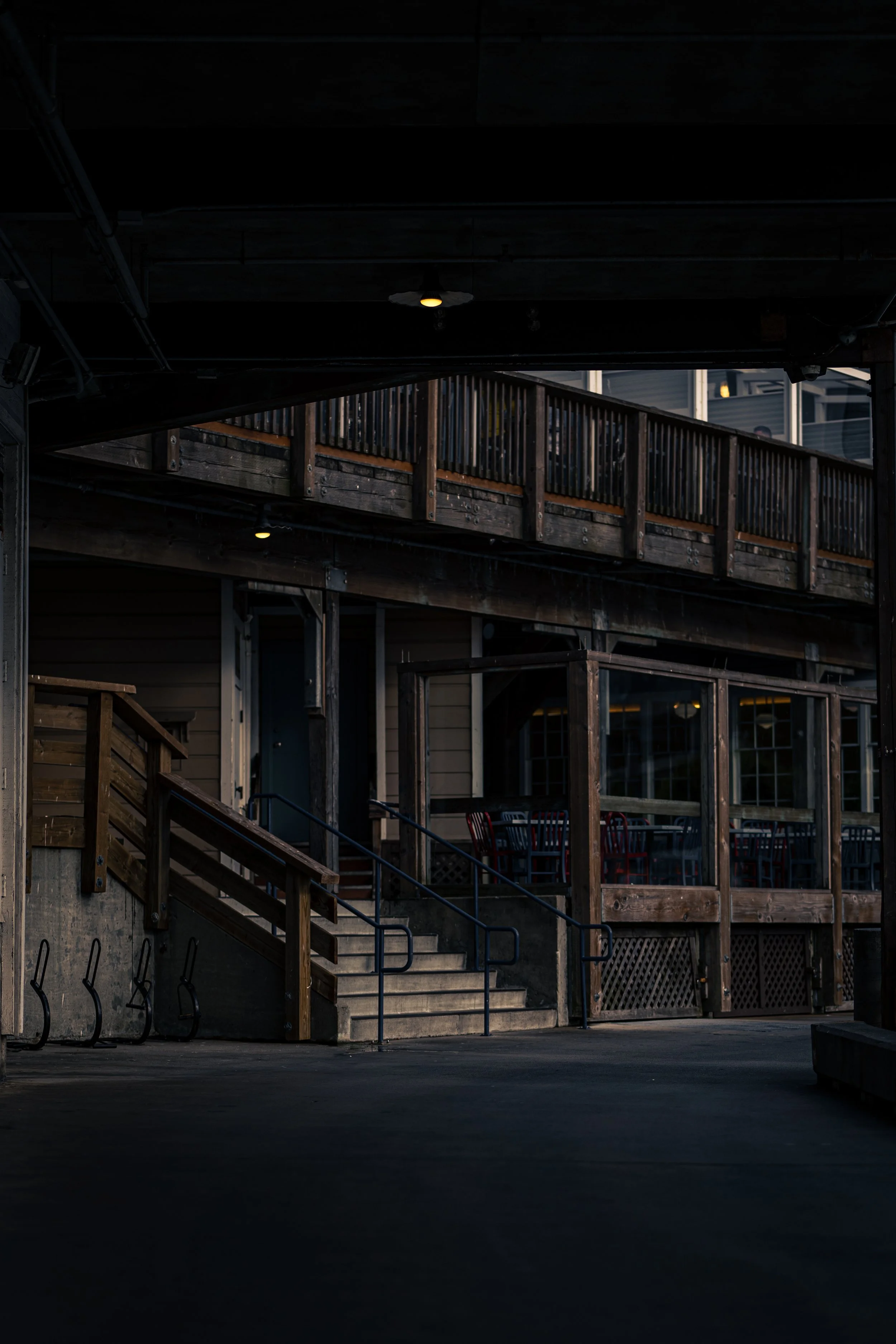 Dimly lit outdoor area with wooden structures, stairs, and railing. Overhead lights and seating visible inside building.