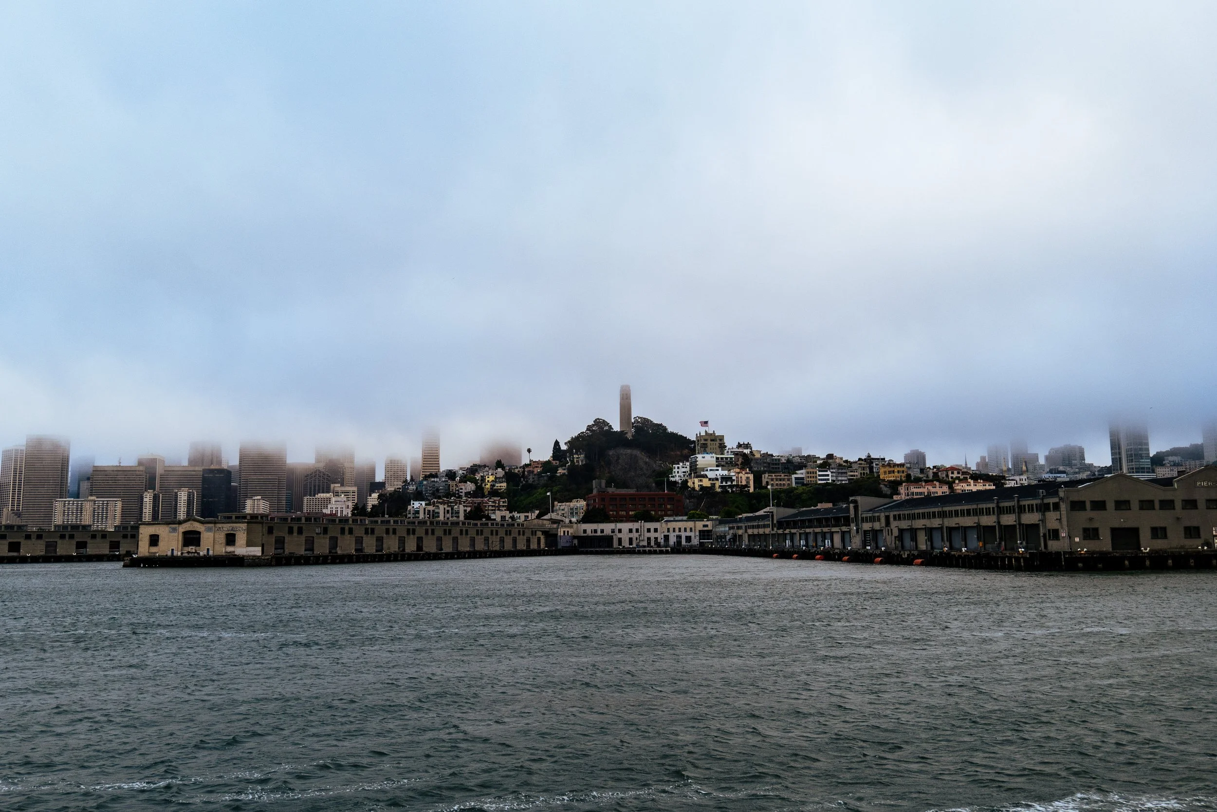Foggy view of San Francisco skyline and waterfront, showcasing Coit Tower and partially obscured buildings.