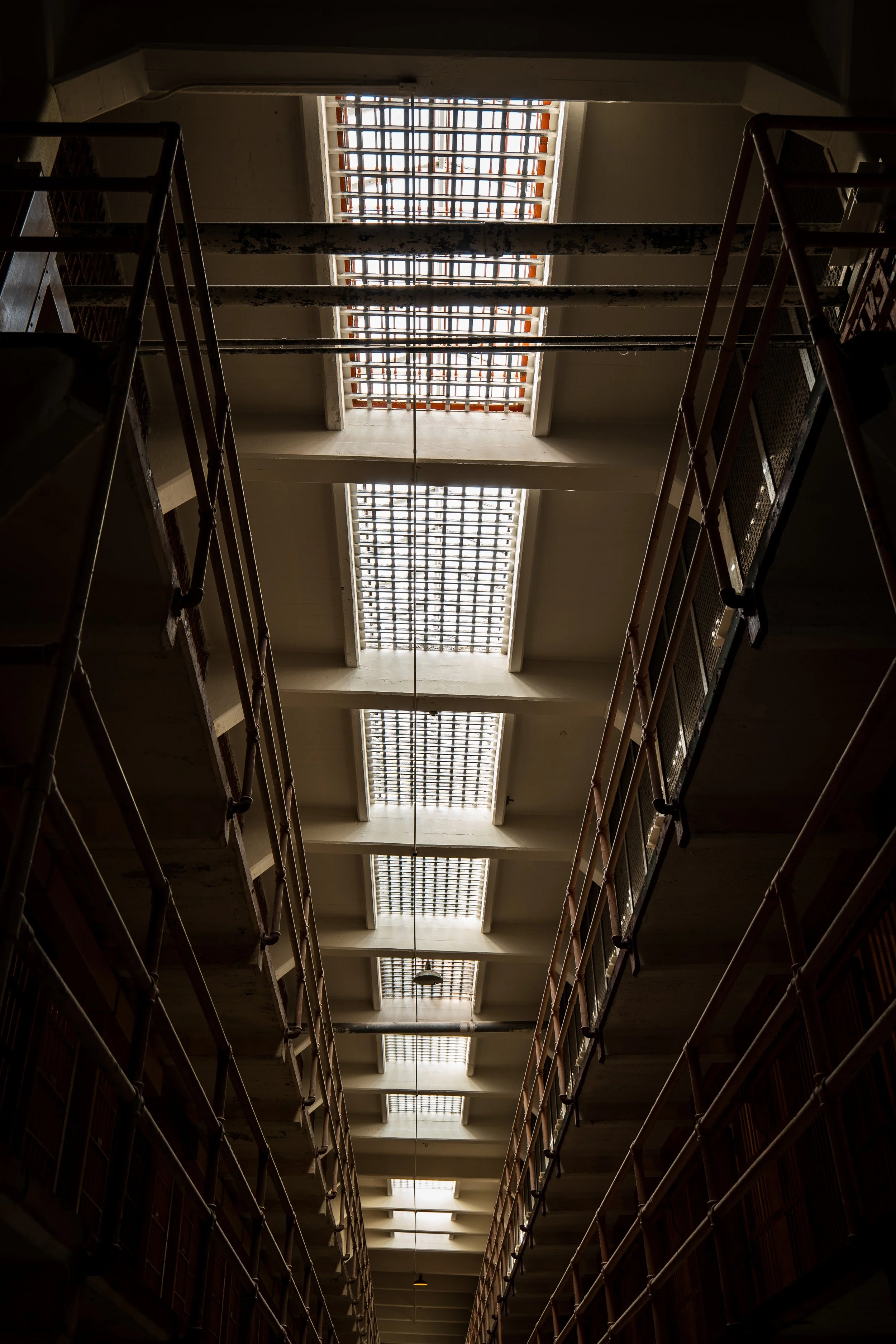View of a prison cell block featuring rows of metal railings and barred windows on the ceiling.