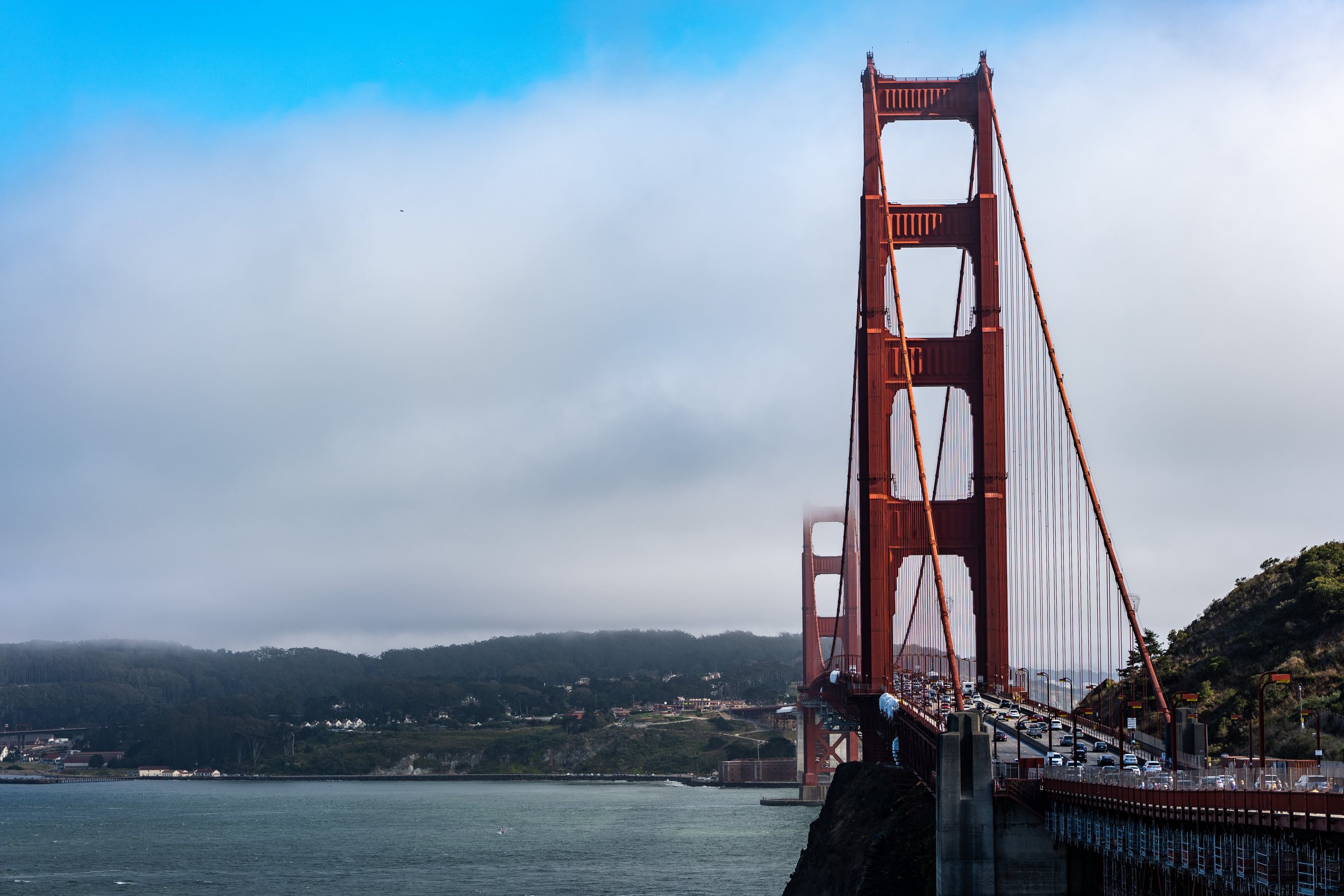 The Golden Gate Bridge extending over water with a cloudy sky and distant hills.