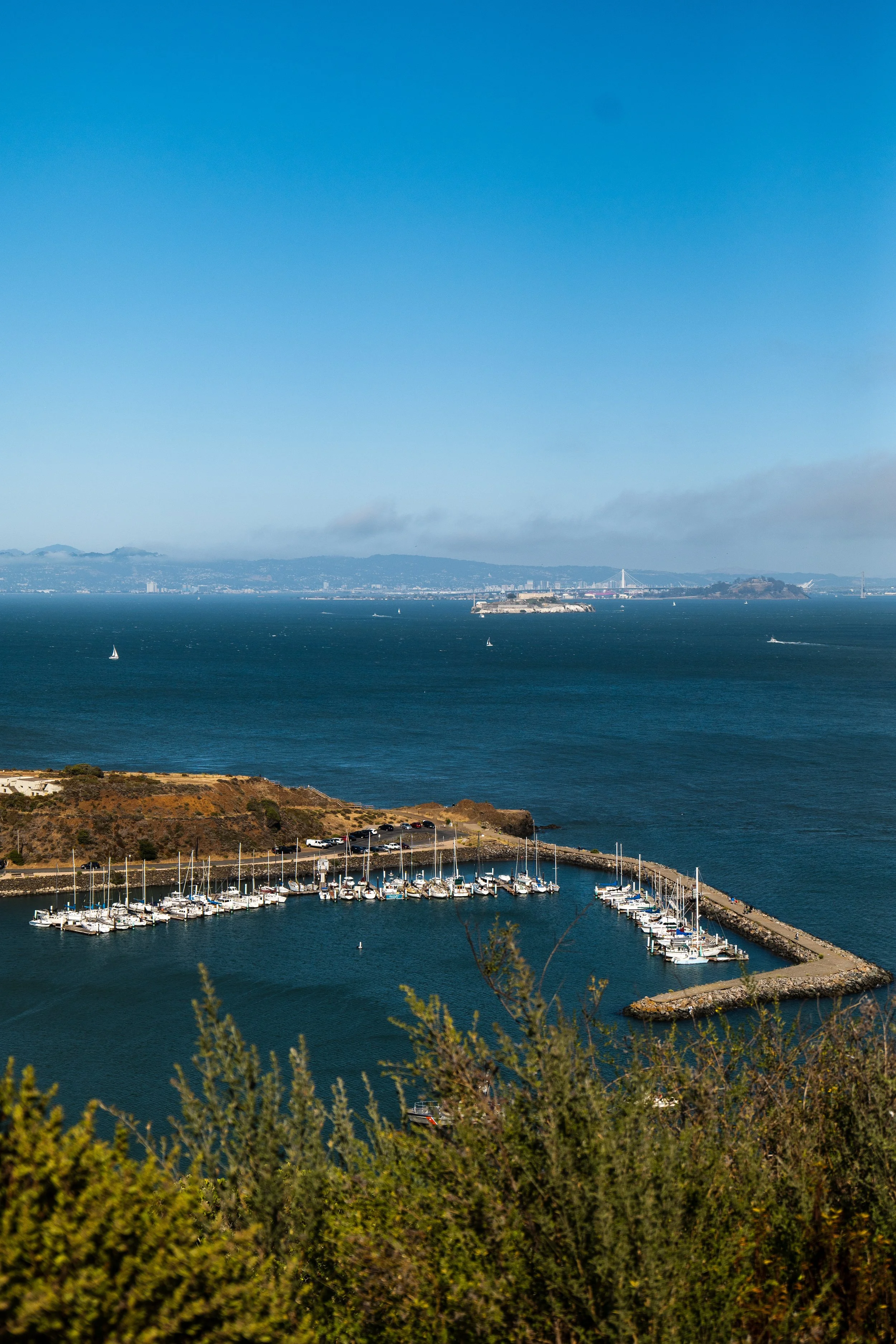 Aerial view of a marina with boats, surrounded by a breakwater, on a large body of water with a distant shoreline and mountains under a clear blue sky.