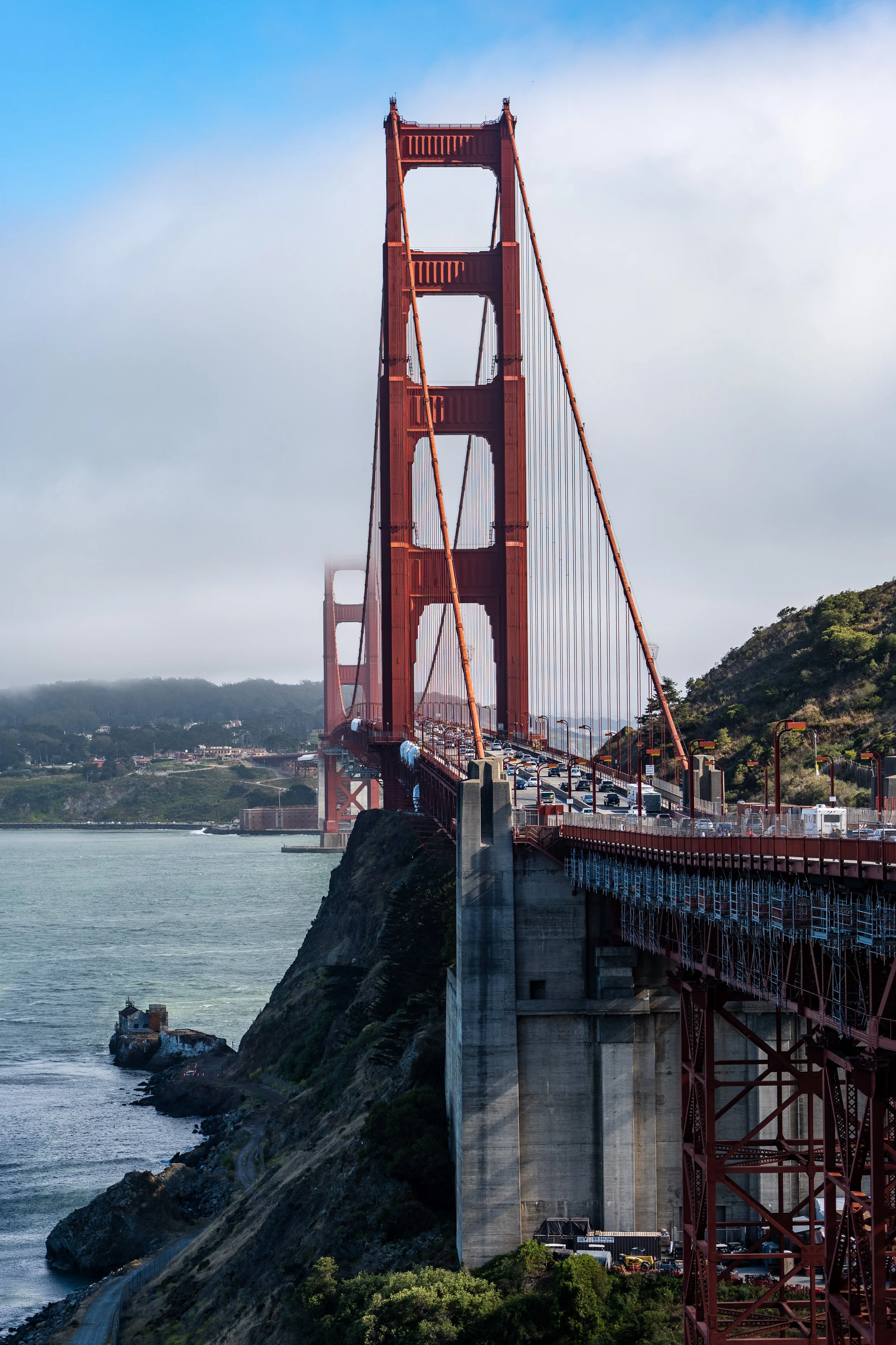 Golden Gate Bridge spanning San Francisco Bay, surrounded by water and rolling hills, with partially cloudy sky.