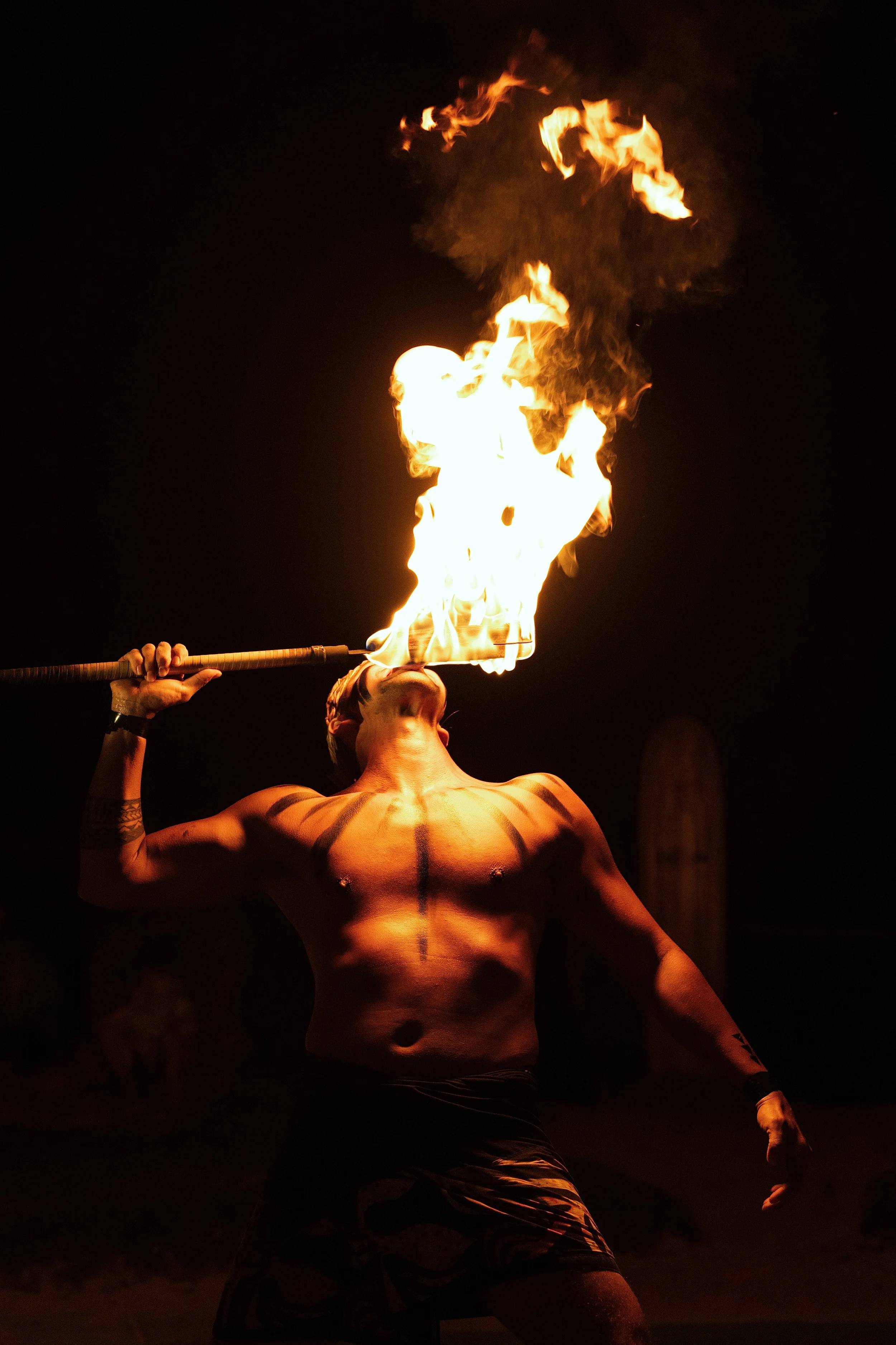 A shirtless person is performing a fire-eating act, holding a flaming torch near their mouth at night, with fire forming a large flame plume.