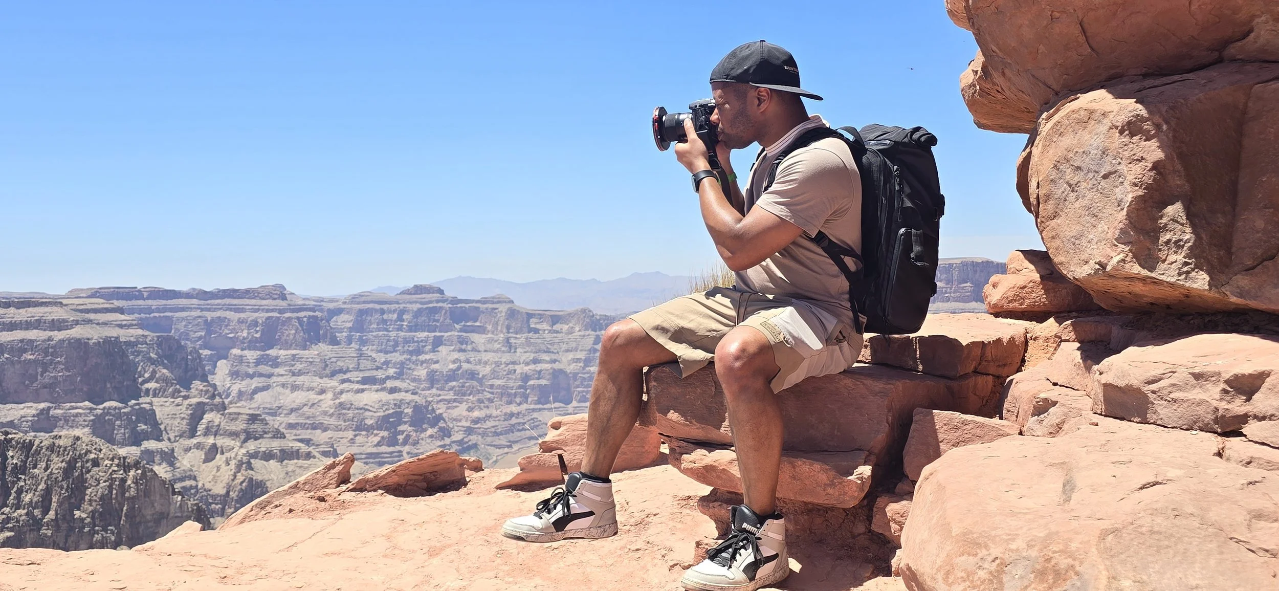 A man sitting on rocks at the Grand Canyon, taking a photograph with a camera in sunny weather.