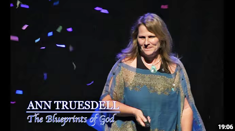 A woman with long hair wearing a blue and gold dress standing on a stage with black background and colorful confetti, with text identifying her as Ann Truesdell and labeled 'The Blueprints of God'.