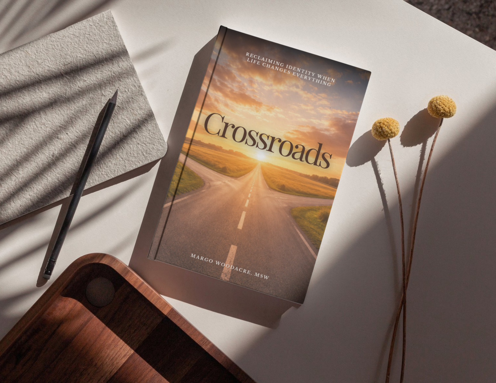 Book titled 'Crossroads' with a cover image of a sunset over a fork in a road, on a white surface next to yellow billy buttons flowers and a notebook with a pen.