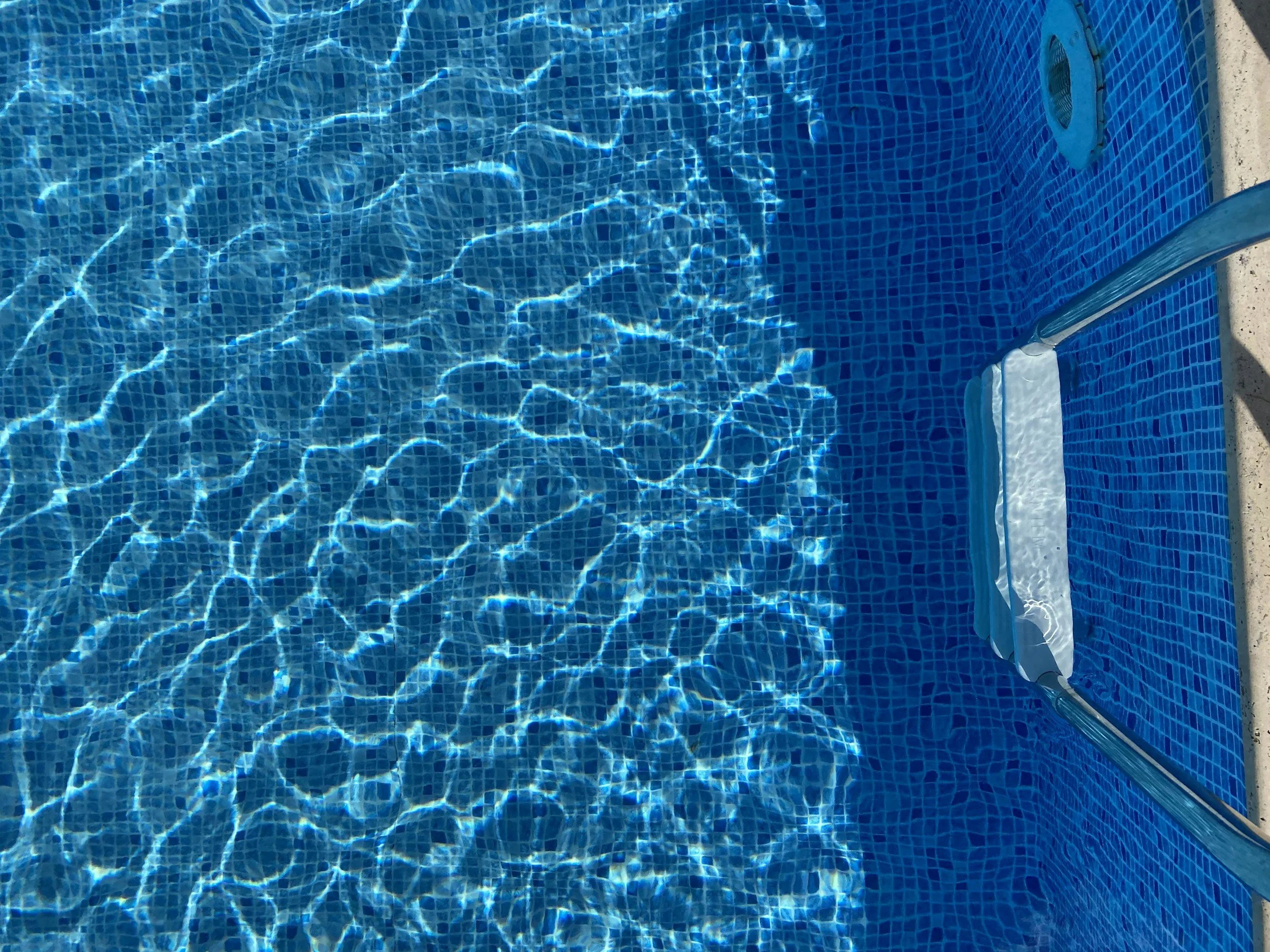 A swimming pool with blue mosaic tiles and clear water, reflecting sunlight. Part of a pool ladder and pool edge are visible.