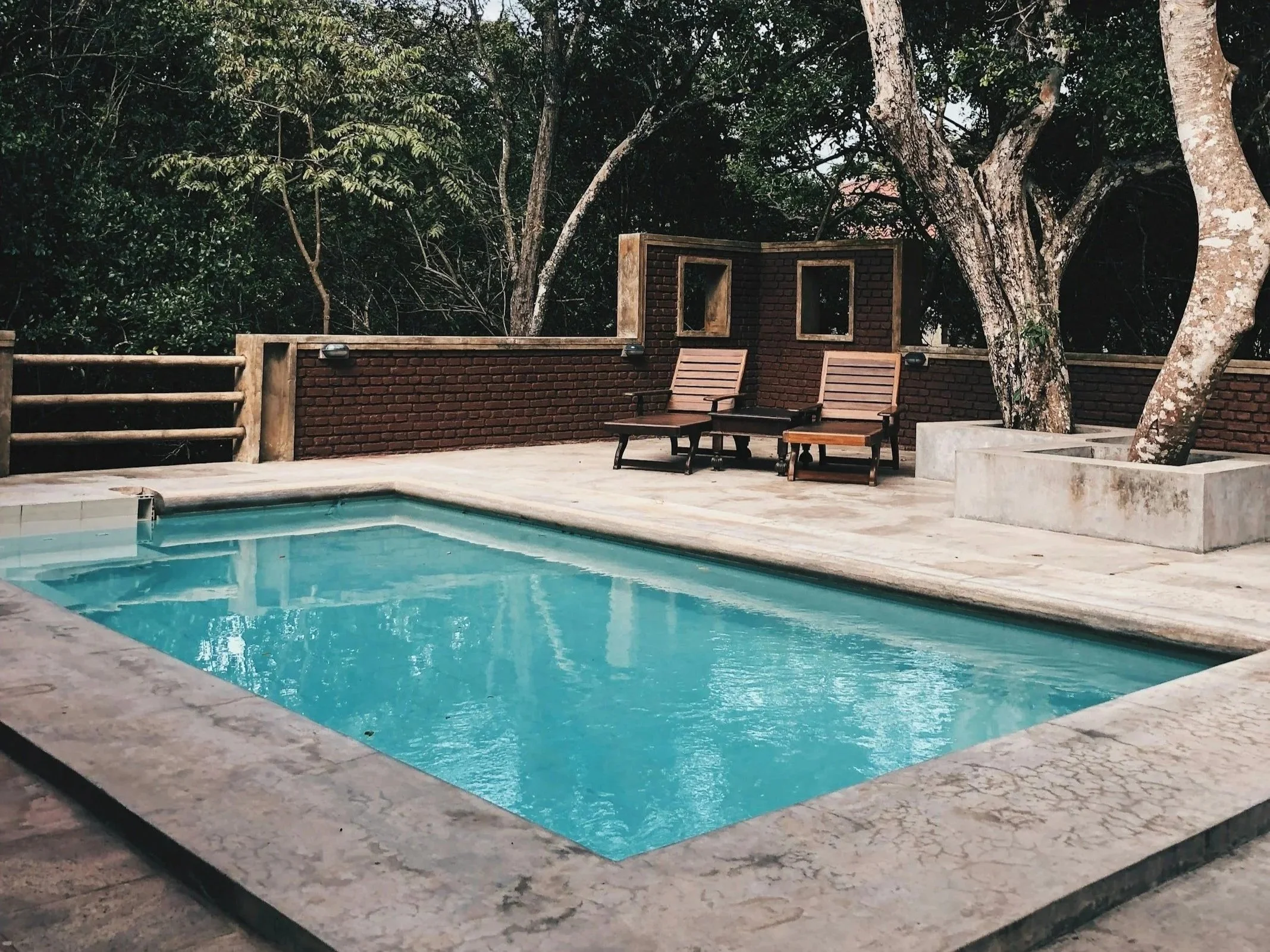 An outdoor swimming pool with clear blue water, surrounded by a stone deck and a brick wall fence. There are wooden chairs and small tables beside the pool, with tall trees and dense greenery in the background.