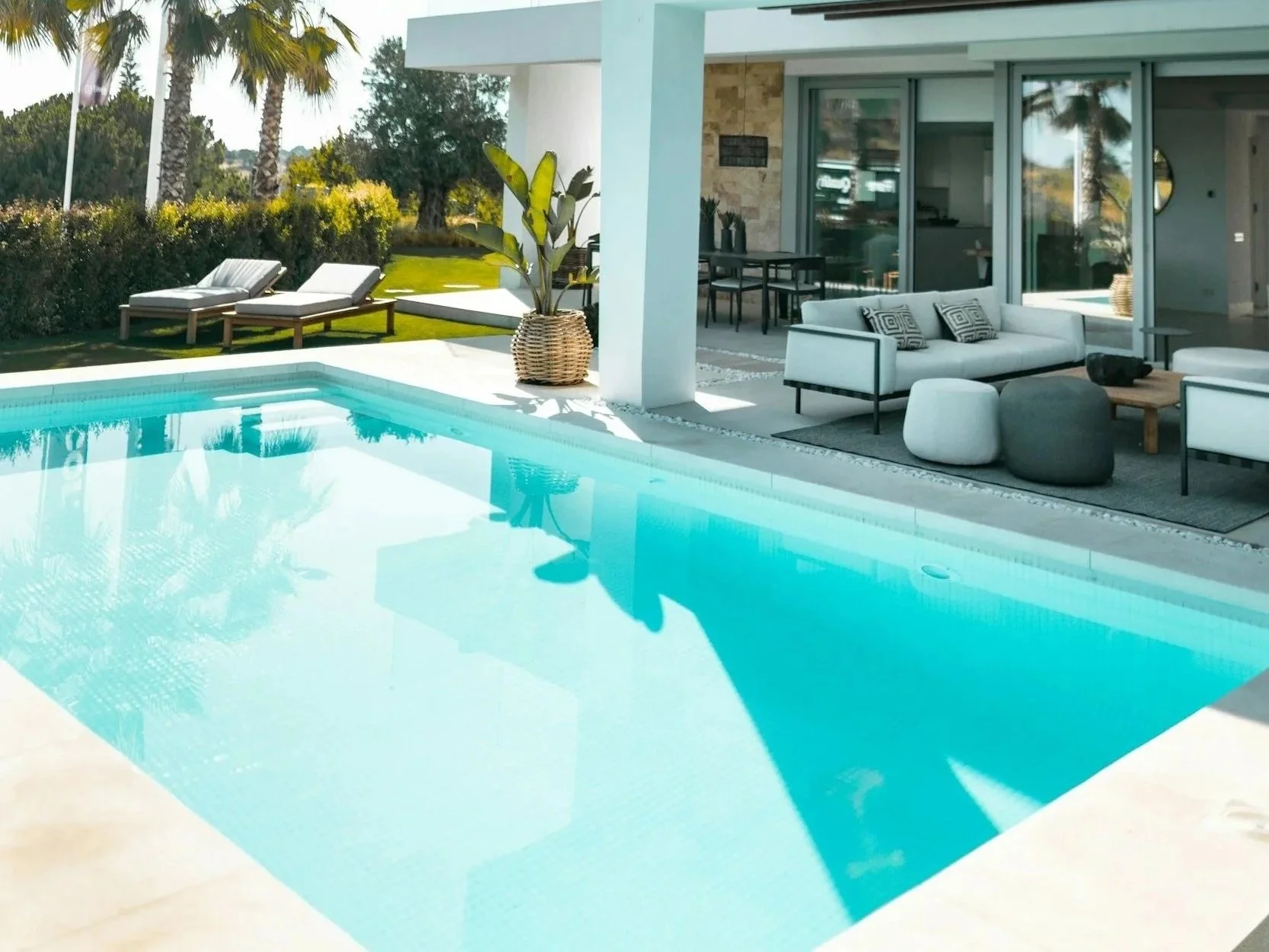 Modern backyard with a swimming pool, outdoor lounge area with white couch and pillows, potted plant, and outdoor chairs under a shaded patio, with green trees and bushes in the background.