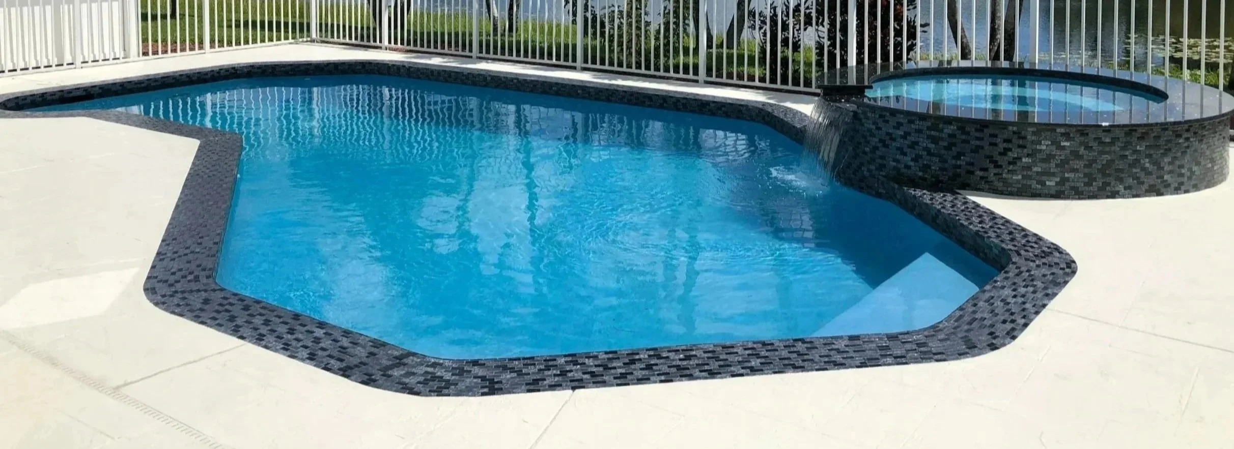 In-ground swimming pool with a spa area, surrounded by light-colored concrete decking, enclosed by a white metal fence, with trees and a mountain view in the background.