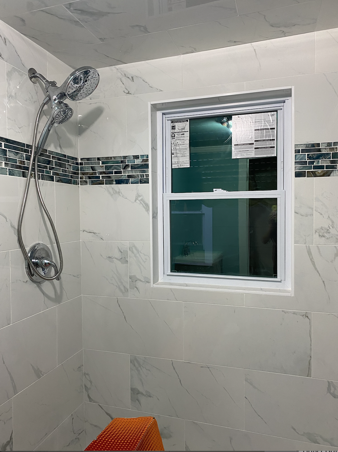 Bathroom shower area with a marble tile wall, a chrome showerhead, window, and a small orange step stool.