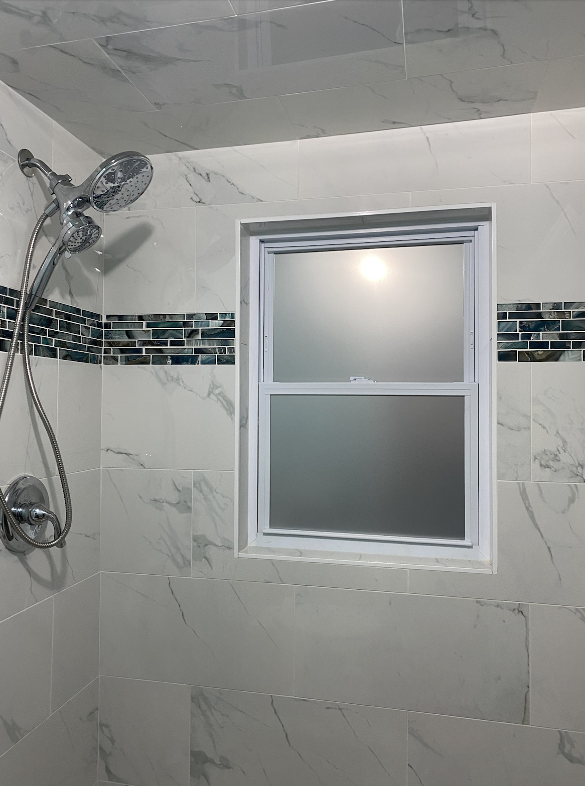 Shower area with a chrome showerhead and a frosted glass window in a bathroom with beige marble tiles and a decorative strip of small, dark-colored tiles.