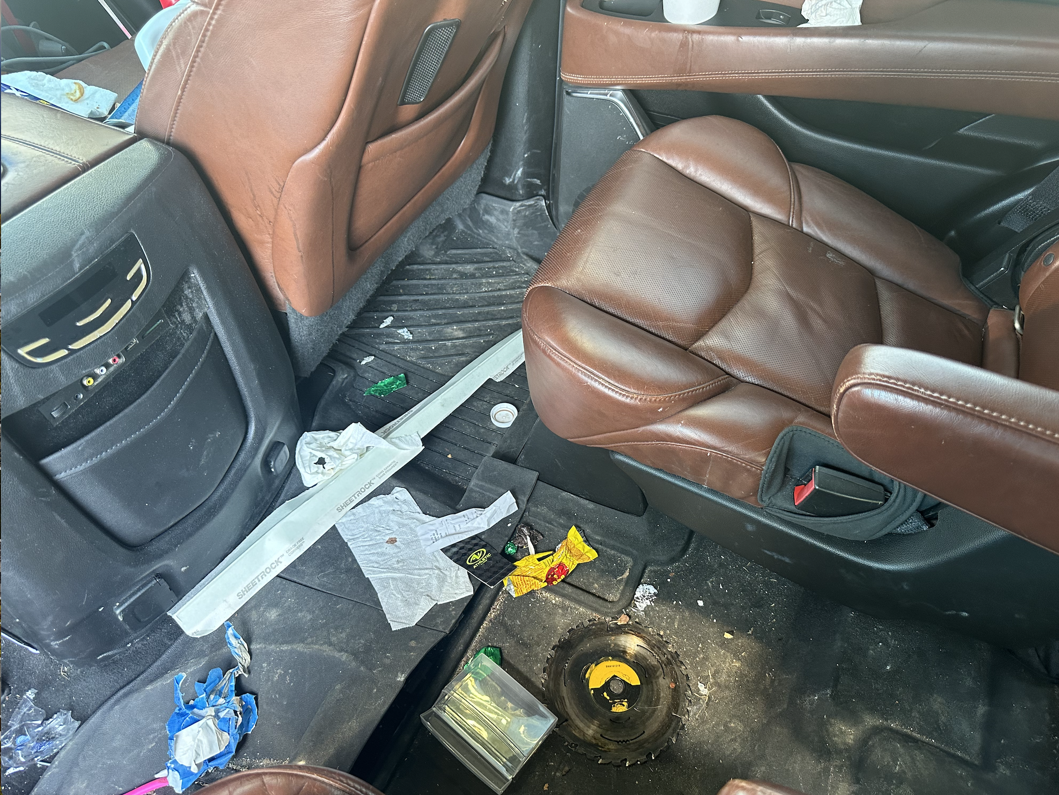 The interior of a car with brown leather seats and black floor mats, showing trash and debris scattered on the floor, including wrappers, tissues, and miscellaneous items.
