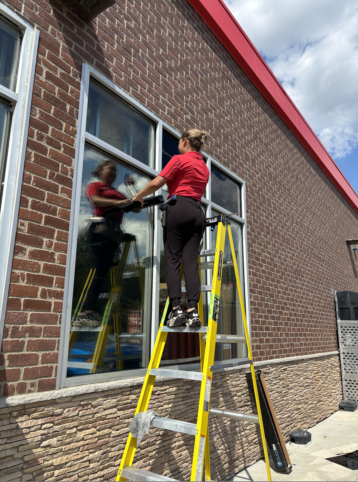 A woman cleaning large glass windows of a brick building while standing on a yellow ladder on a sunny day.