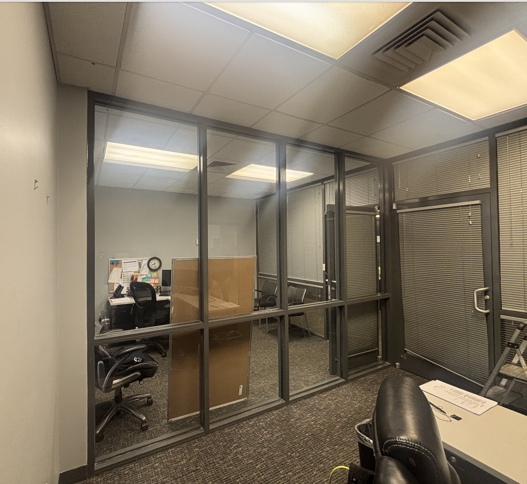 An office conference room with a glass wall and blinds, featuring chairs, a table, and a bulletin board by the wall.