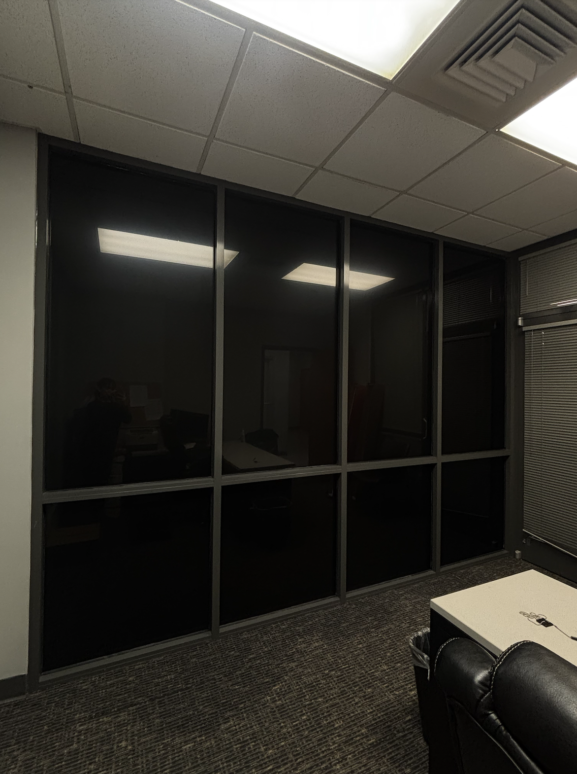 An office viewed through a glass partition with black frames, showing a person at a desk and fluorescent ceiling lights, with blinds on the windows to the right.