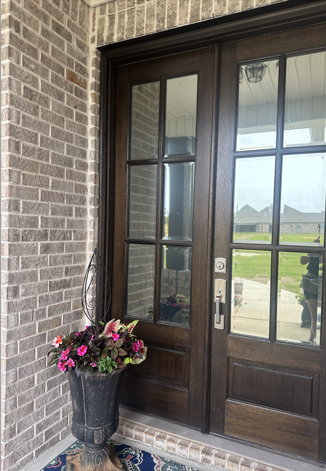 Front door with glass panes, brick exterior wall, a potted plant with pink flowers, and a porch rug.