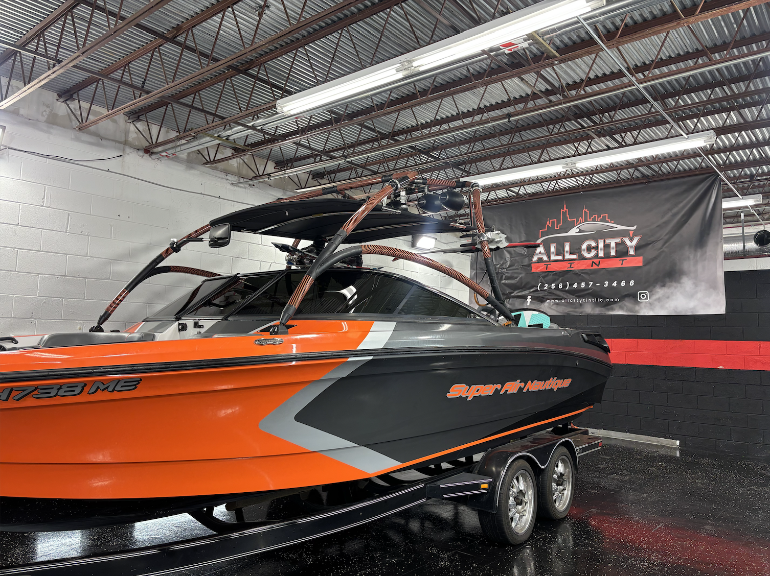 A speedboat with orange, black, and gray colors on a trailer inside a garage. The boat has a sleek design with the words 'Super Air Nautique' on the side and registration number 'AL7388 ME'. A banner on the wall reads 'All City TIN' with contact info