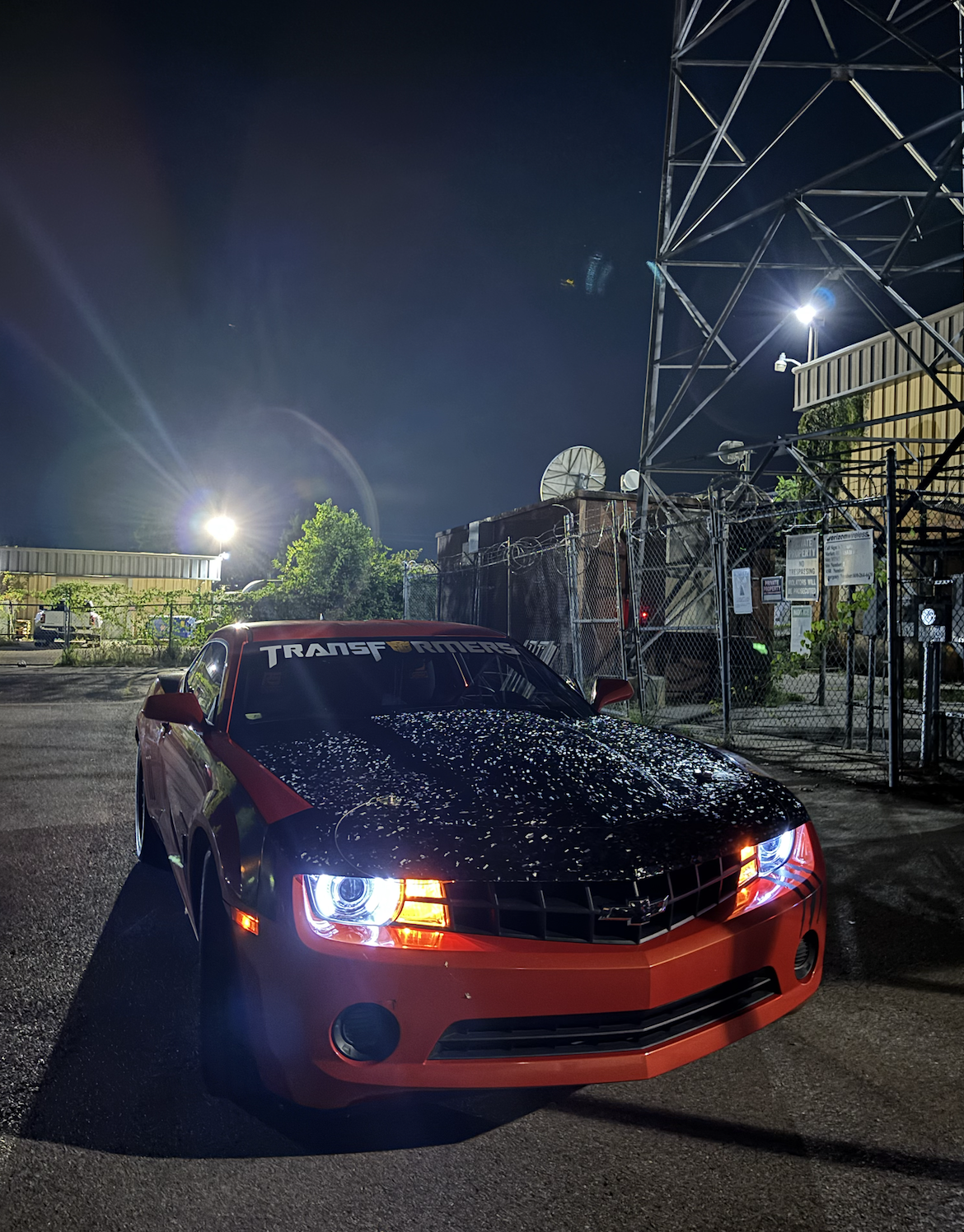 Red and black Chevrolet Camaro with Transfomers sticker on the windshield, parked outdoors at night under bright lights, with a chain-link fence and industrial structures in the background.