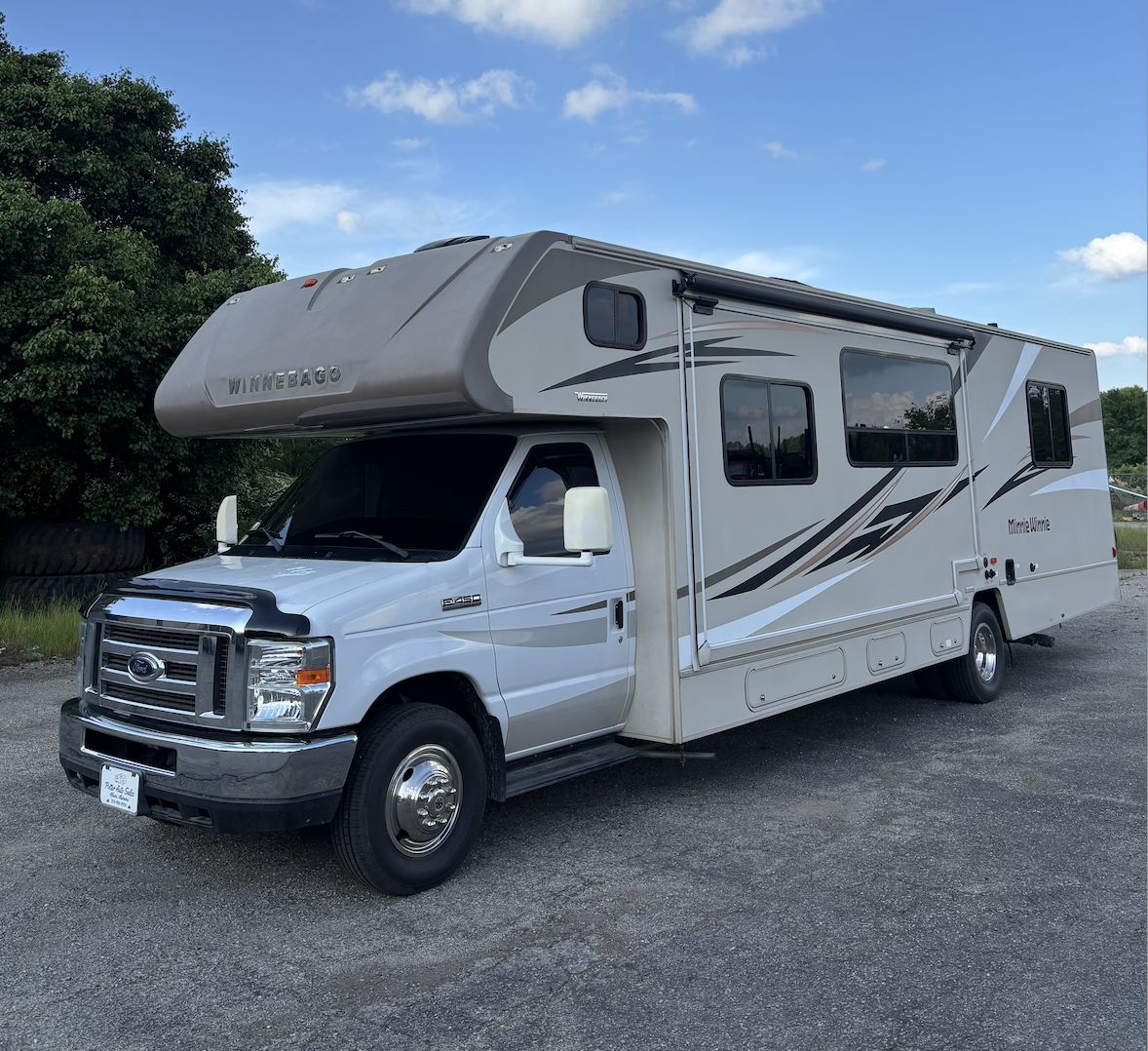 A white and gray Winnebago motorhome parked on a gravel surface under a partly cloudy sky.