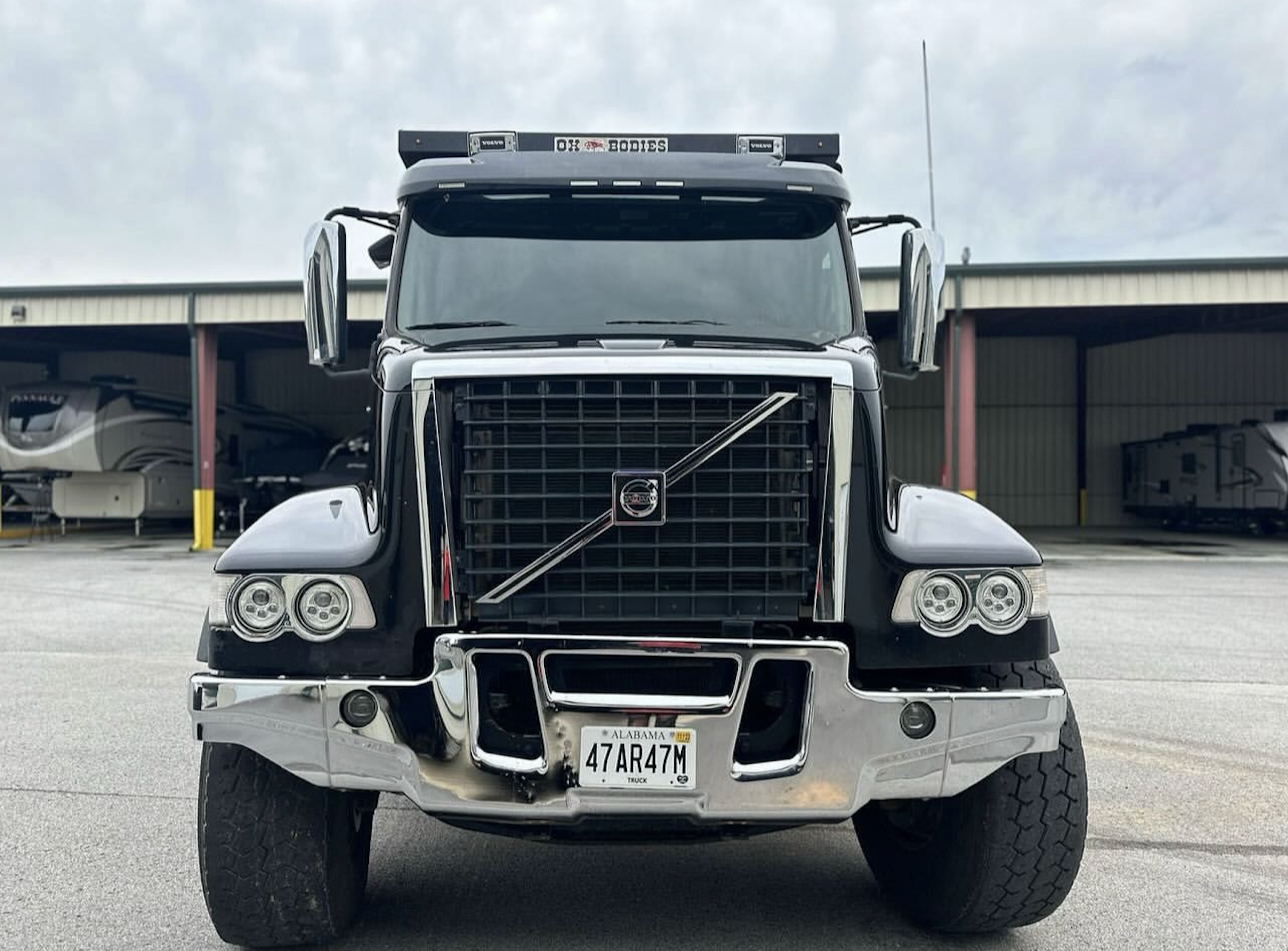 Front view of a black semi-truck with a chrome grille, large side mirrors, and Alabama license plate, parked in an outdoor lot with RVs in the background.
