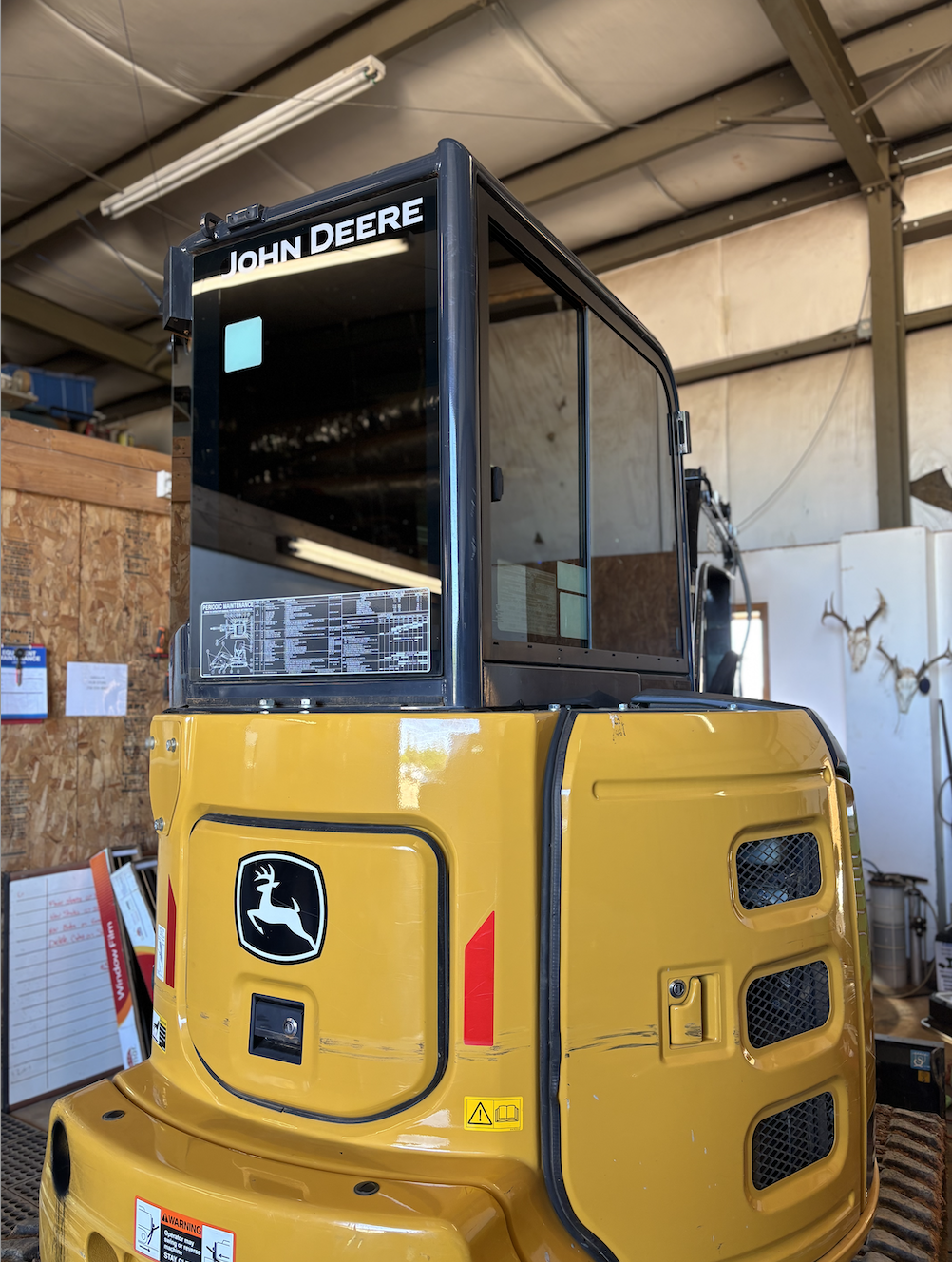 A yellow John Deere compact excavator positioned inside a workshop or warehouse. The black upper section features the John Deere logo and a sign reading 'JOHN DEERE'. The workshop has a wooden wall, some equipment, and a whiteboard in the background.
