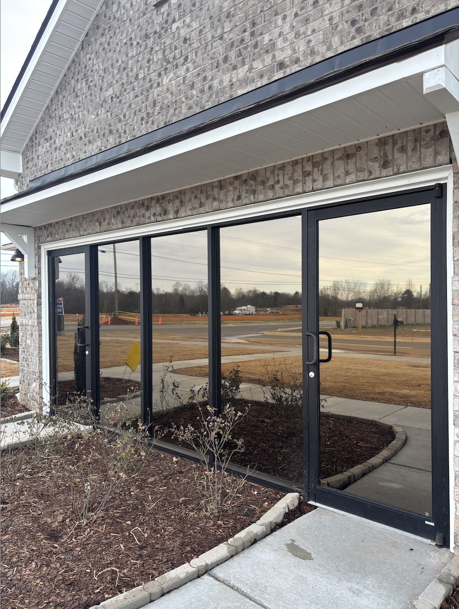  storefront with large glass windows and door, brick exterior, small landscaped area with plants, sidewalk, street with cars and trees in background.
