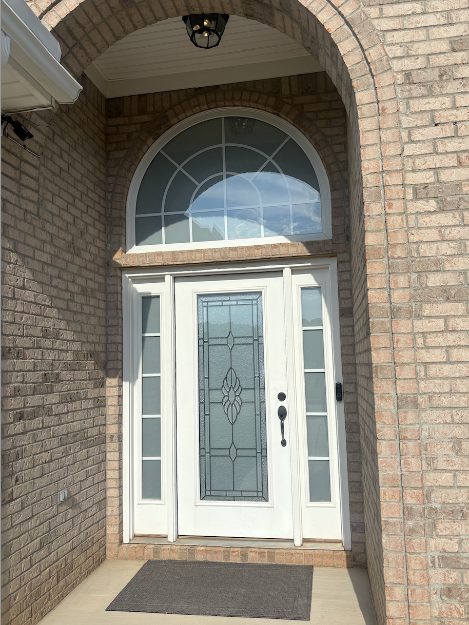 Front door with glass inset, flanked by sidelights, above a semi-circular window with geometric windowpanes, set in a brick wall.