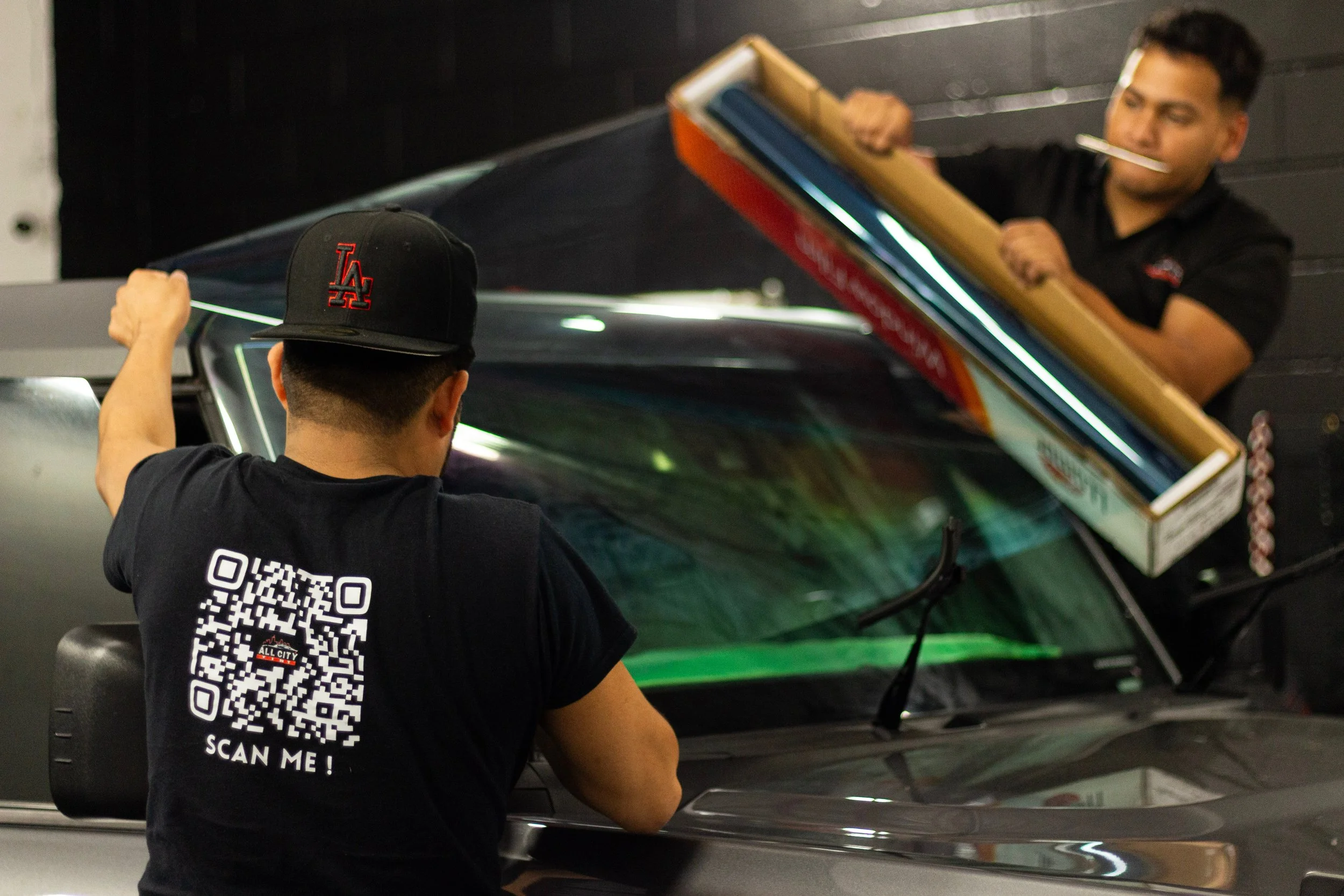 Man in black shirt with QR code on back, sitting in front of a car, with another man standing on the other side of the car holding a box.
