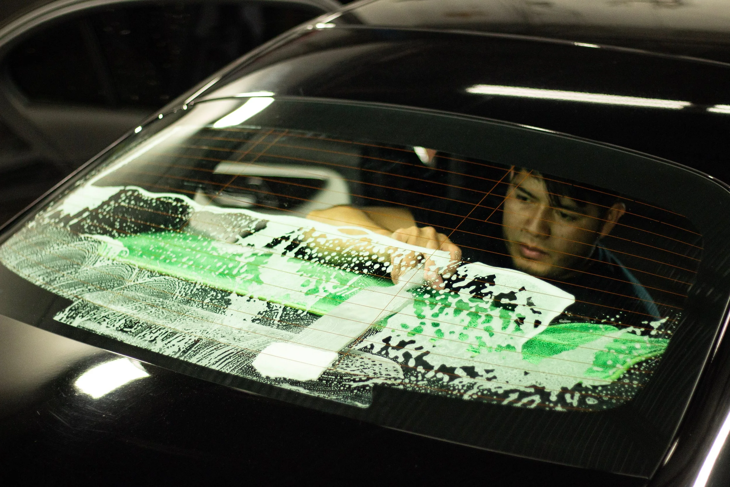 A person cleaning the rear window of a black car with soap and water, with a portion of the person's face visible through the wet glass.