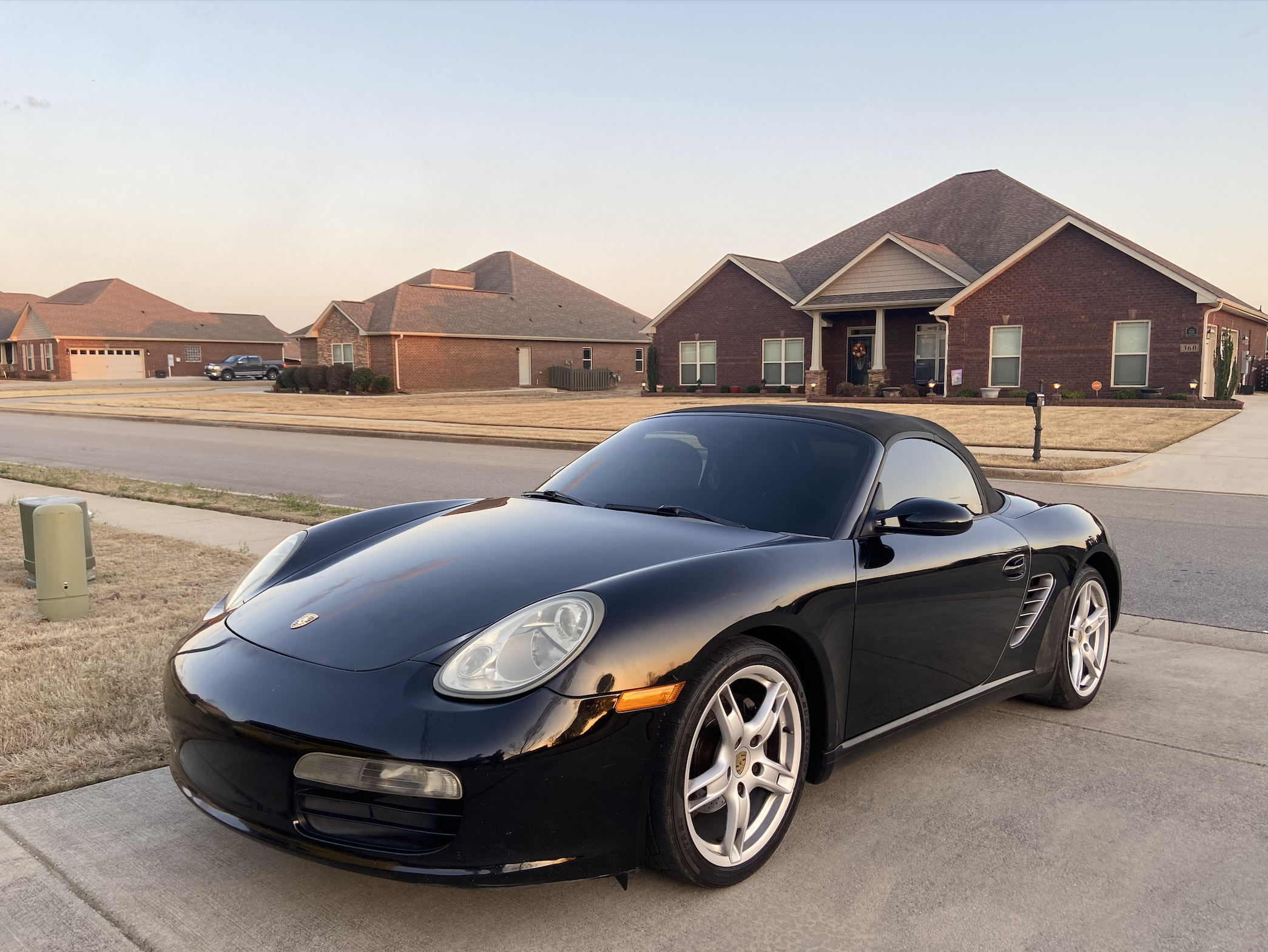 A black Porsche sports car parked on a suburban driveway in front of brick houses under a clear sky.