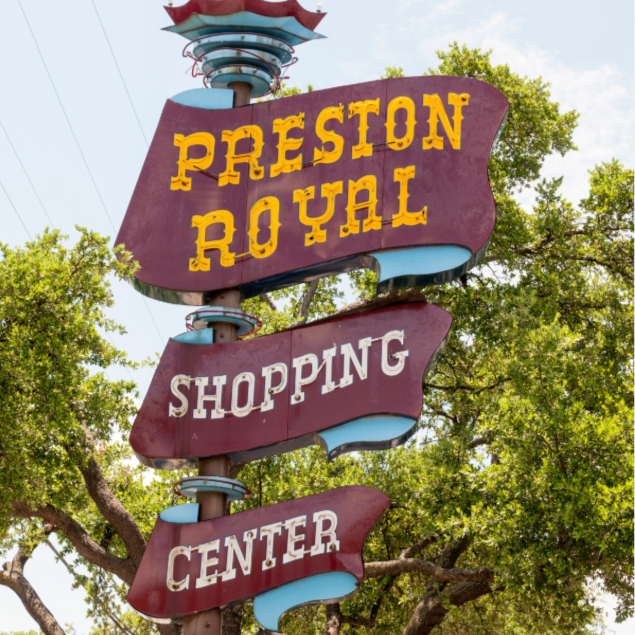 Sign for Preston Royal Shopping Center with yellow and white text on purple banners, set against a background of trees and a partly cloudy sky.