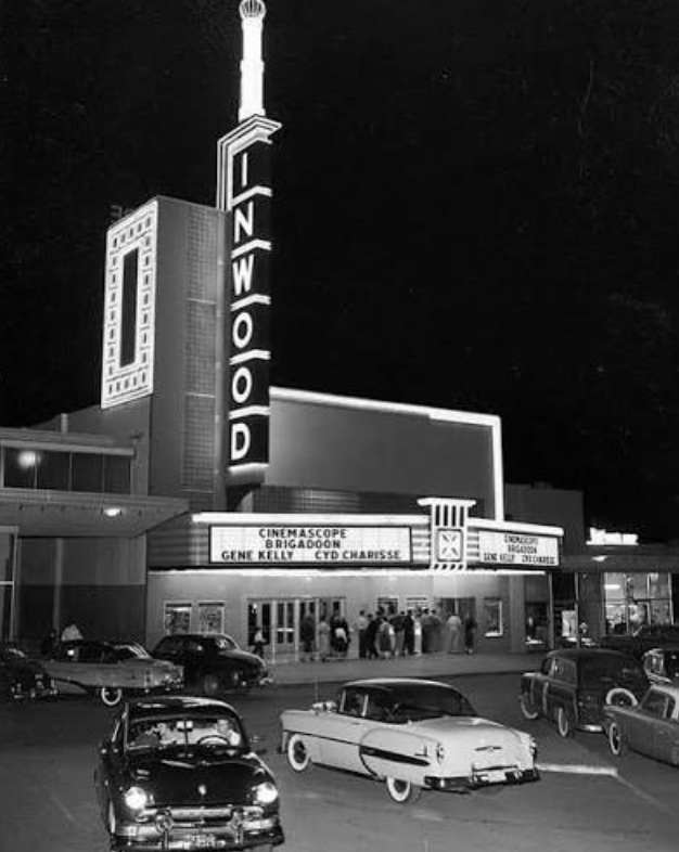 Vintage movie theater at night with neon sign reading 'Hollywood' and a marquee advertising films, with classic cars parked in front.