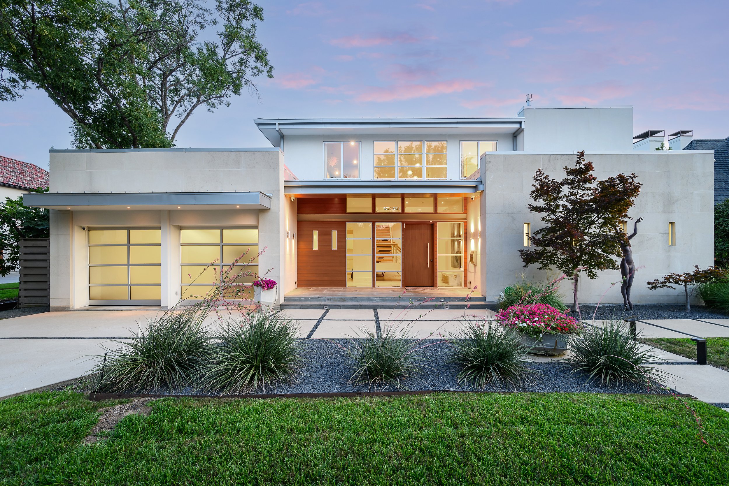 Modern two-story house with large windows, a tree in the front yard, and well-maintained landscaping, including green grass and flowering plants, during dusk.