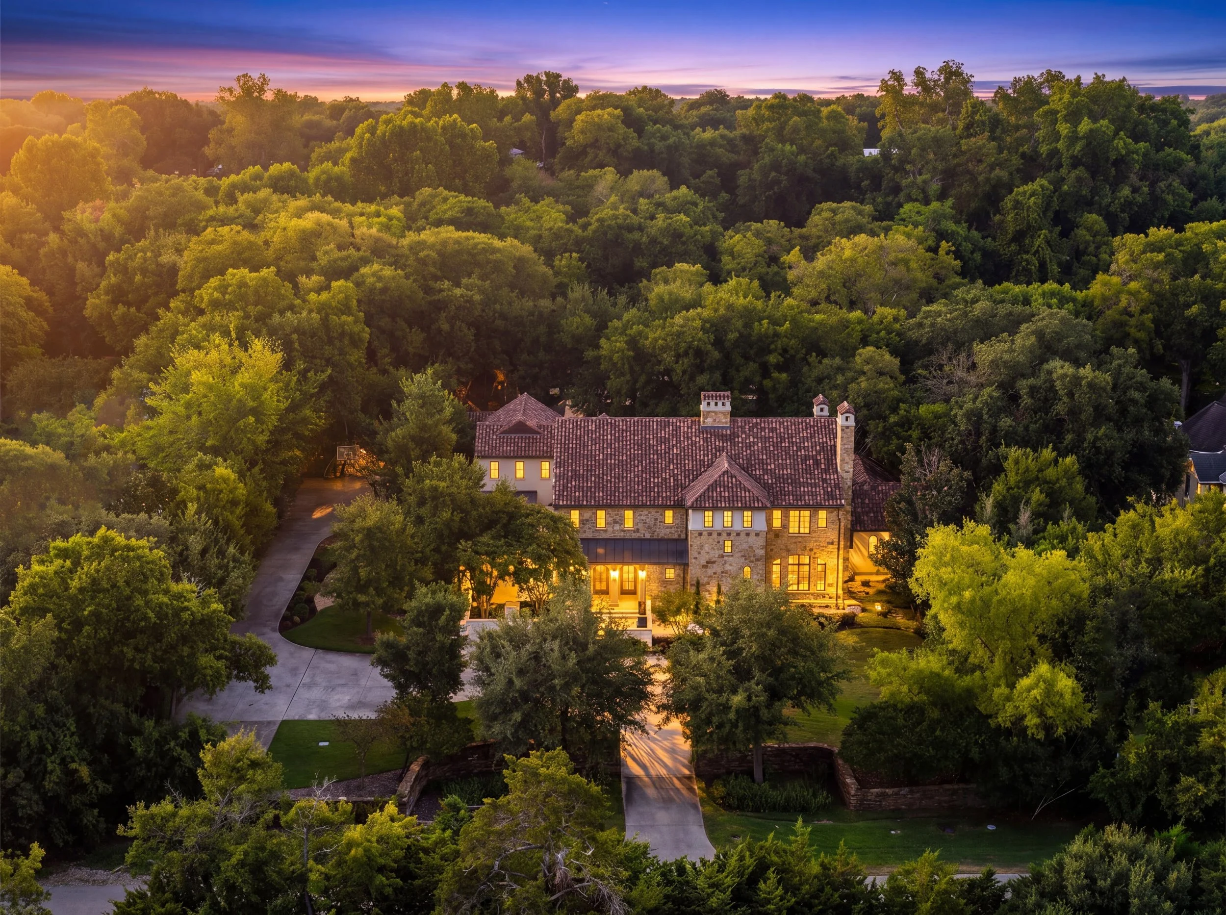 A large house with a brick and stone exterior, red tile roof, and tall chimney, surrounded by dense green trees, under a colorful sunset sky.