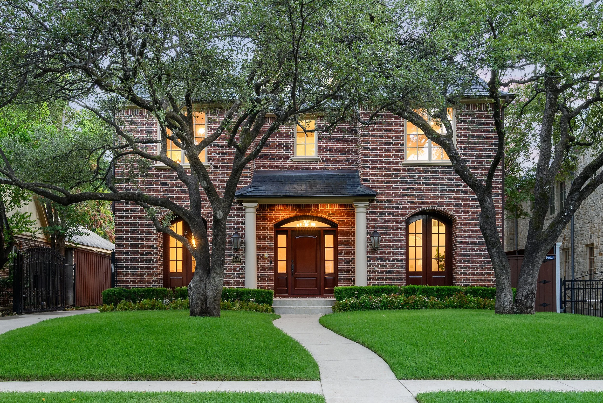 A brick house with large windows and a front door, surrounded by green grass and trees with dense foliage, during early evening.