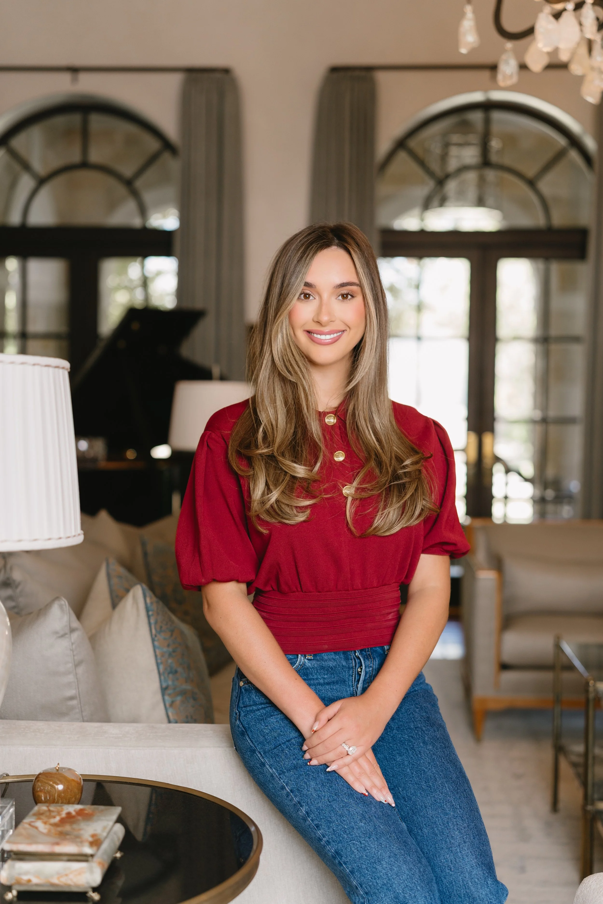 A young woman with long, wavy blonde hair, smiling, wearing a red blouse and blue jeans, sitting on a sofa in a well-lit, elegant living room with large windows and drapes, with a piano and chandelier in the background.