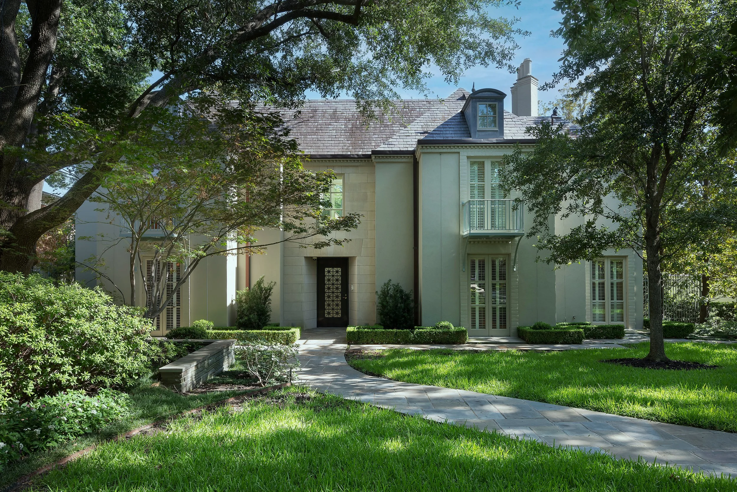 Front view of a beige house with a slate roof, surrounded by lush green trees and a well-maintained lawn, with a sidewalk leading to the front door.