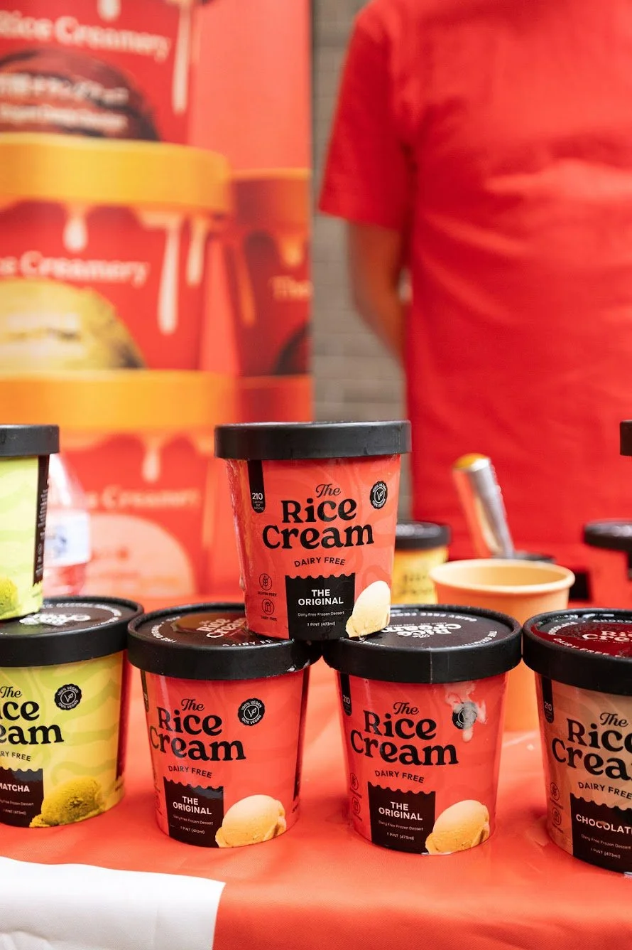 Display of dairy-free rice cream desserts with black lids on a table, with three types visible: original, matcha, and chocolate, in front of a large poster.